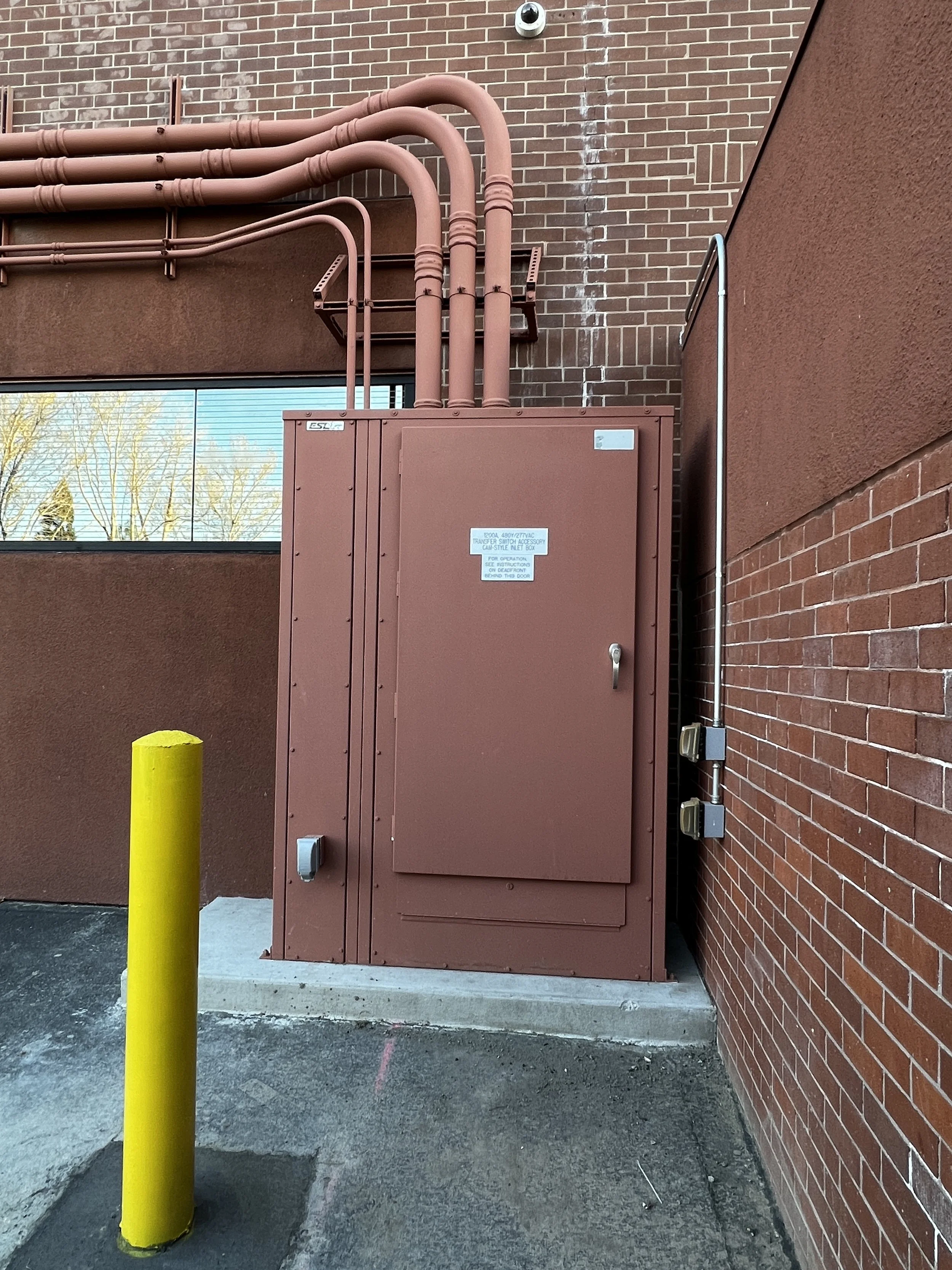Electrical transformer box outside a building with security camera and yellow safety pole.