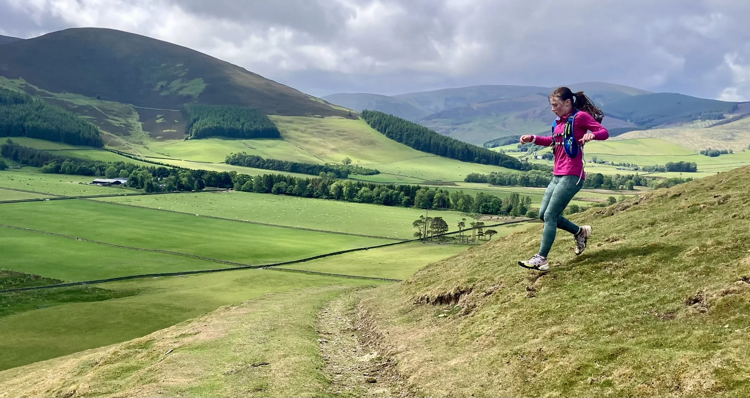 A young woman wearing a pink jacket, gray leggings, and hiking shoes descending a grassy hillside trail with green rolling fields, trees, and mountains in the background under a partly cloudy sky.