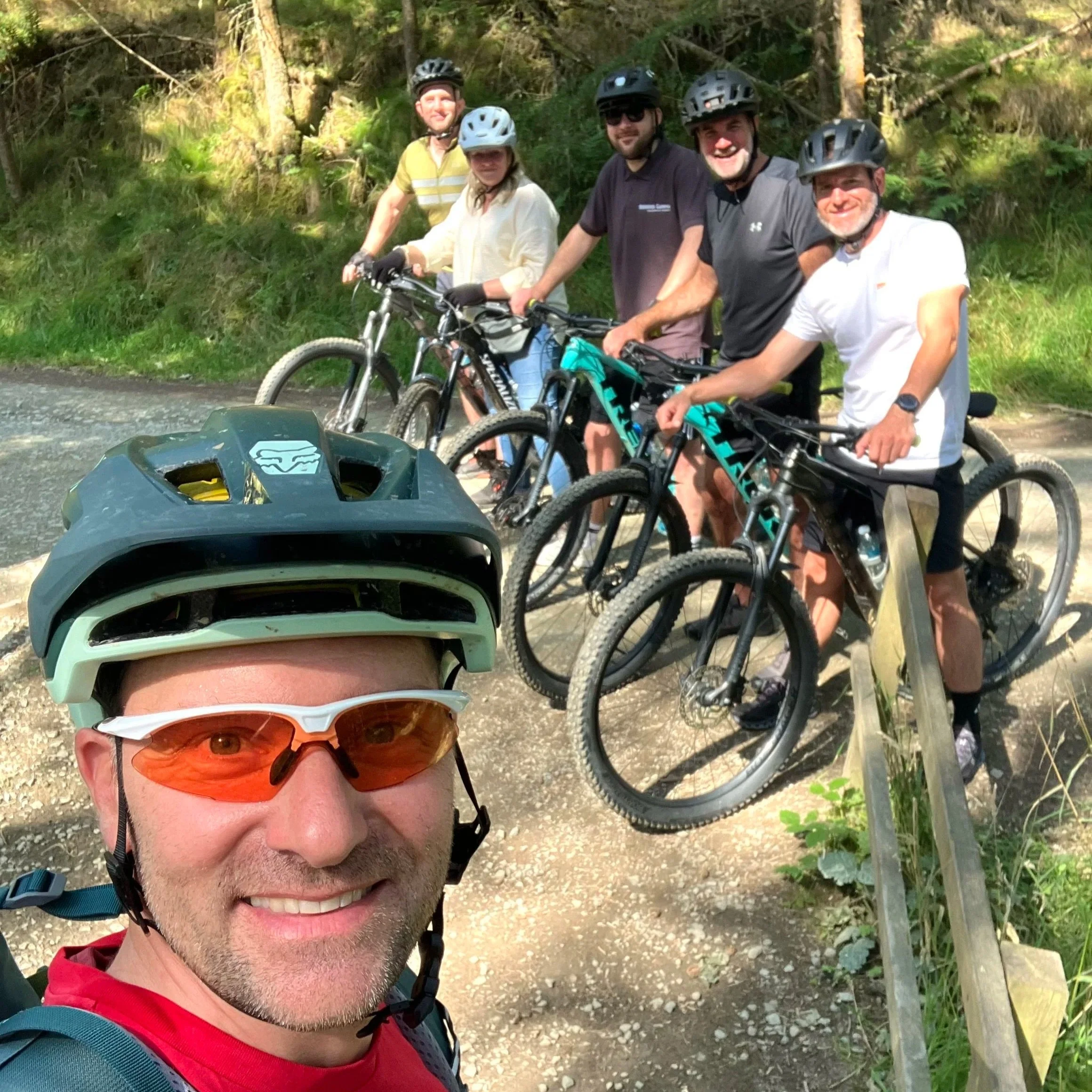Group of six people with mountain bikes outdoors on a trail, wearing helmets and sunglasses, smiling for a selfie taken by one person in the front, with lush green trees in the background.