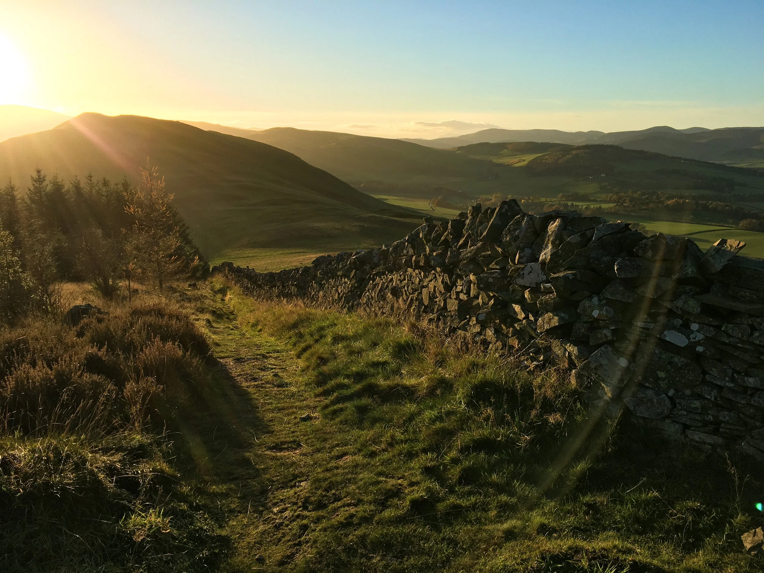 Sunset over rolling green hills with a stone wall and a grassy path in the foreground.