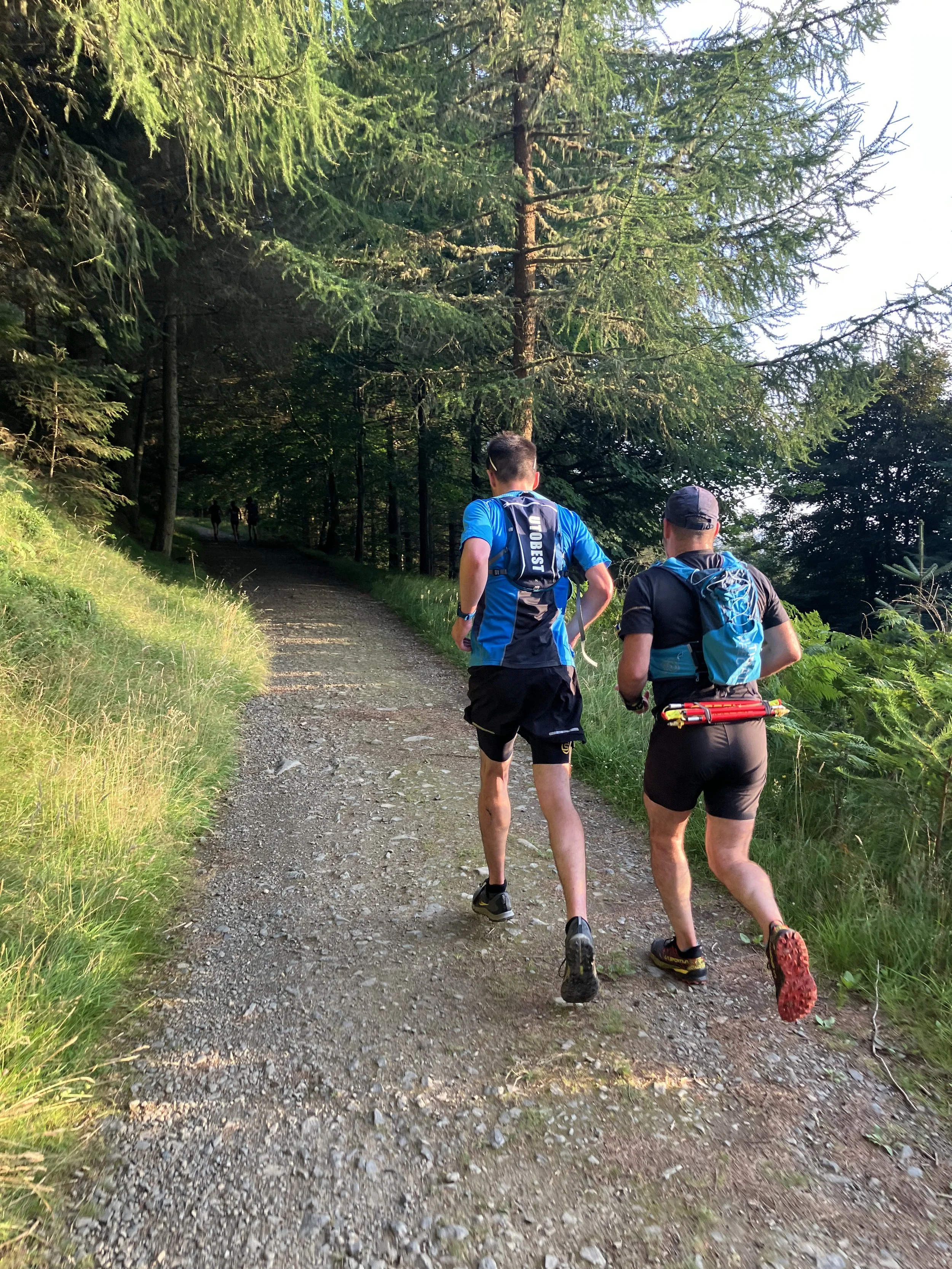Two men running on a forest trail with trees and greenery around.