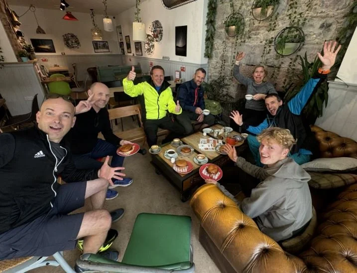 Group of seven friends enjoying coffee and pastries in a cozy cafe, smiling and waving at the camera.