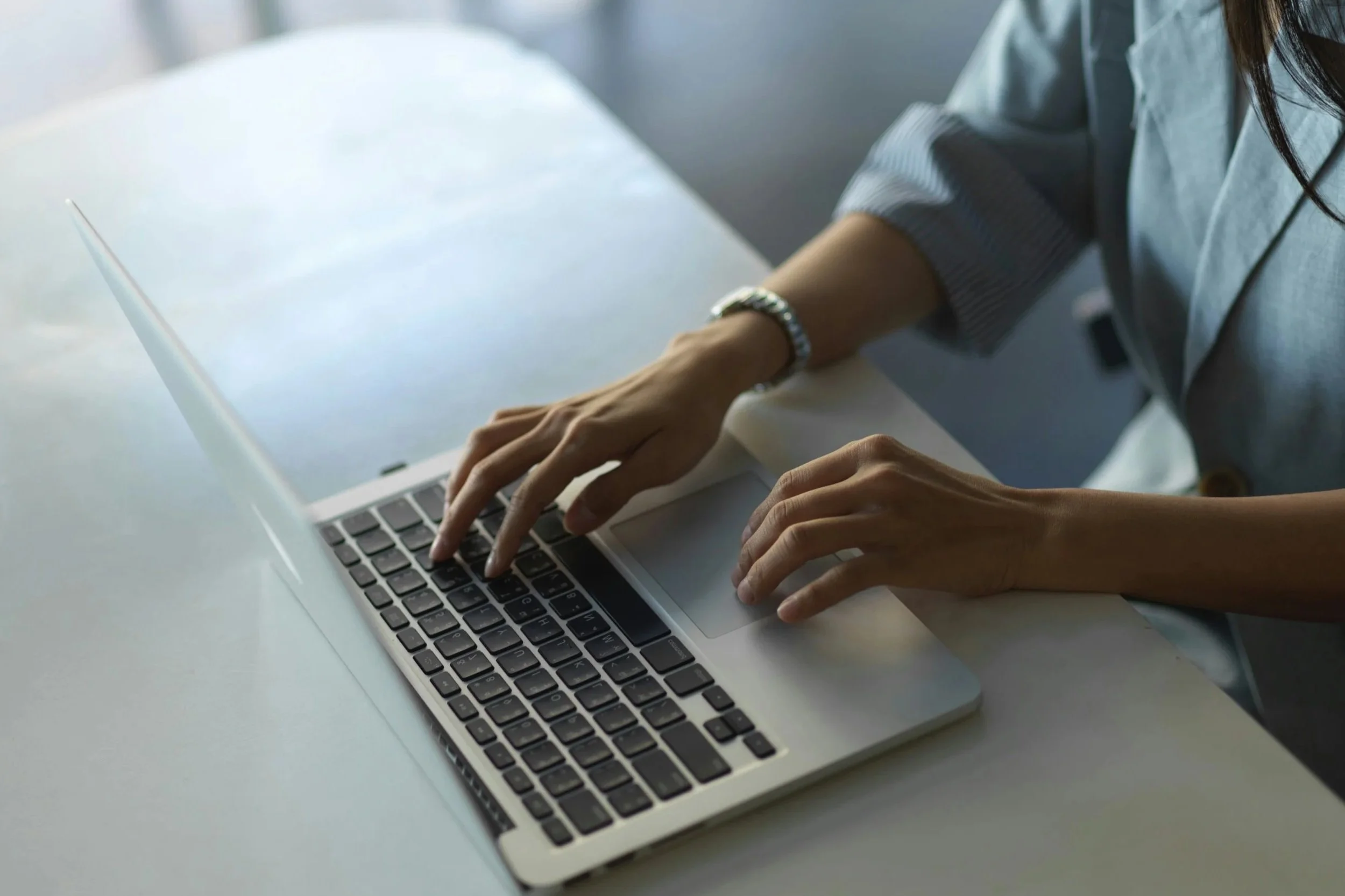Person typing on a laptop keyboard at a white table, wearing a watch and a gray long-sleeve shirt.