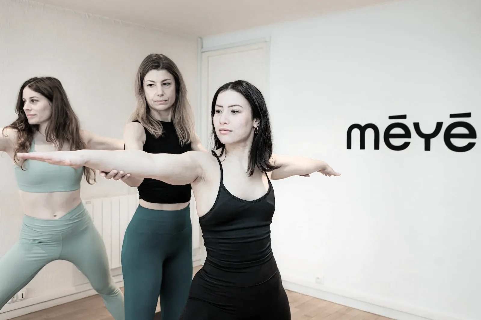 Trois femmes pratiquent le yoga ou la méditation dans une salle blanche, avec le mot 'mèyé' écrit au mur.