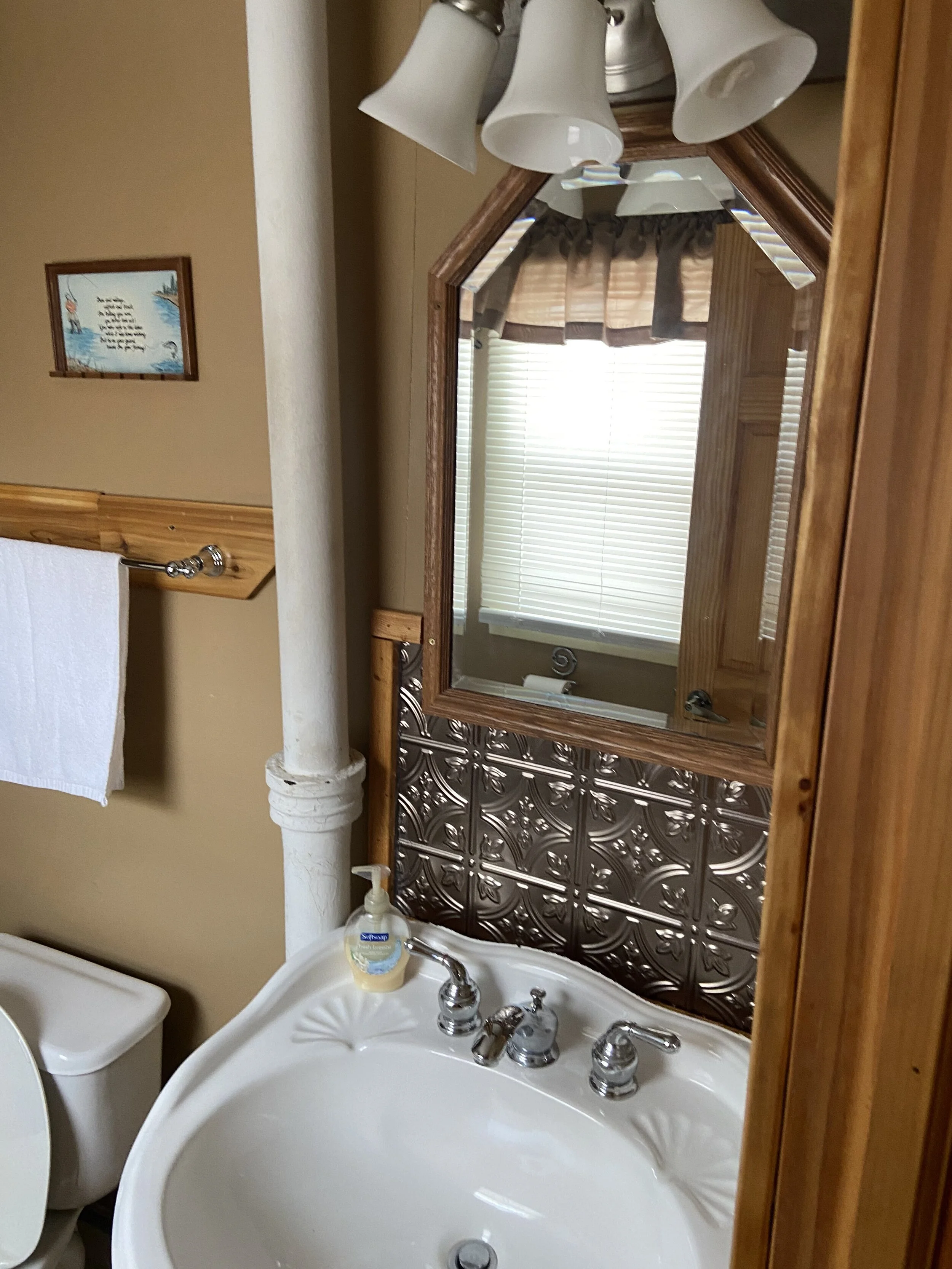 A bathroom with a white sink, a mirror, a wooden cabinet, a white toilet, a towel bar, and a small window with blinds and a valance. The sink has a liquid soap dispenser, and above it is a light fixture with multiple white glass shades.
