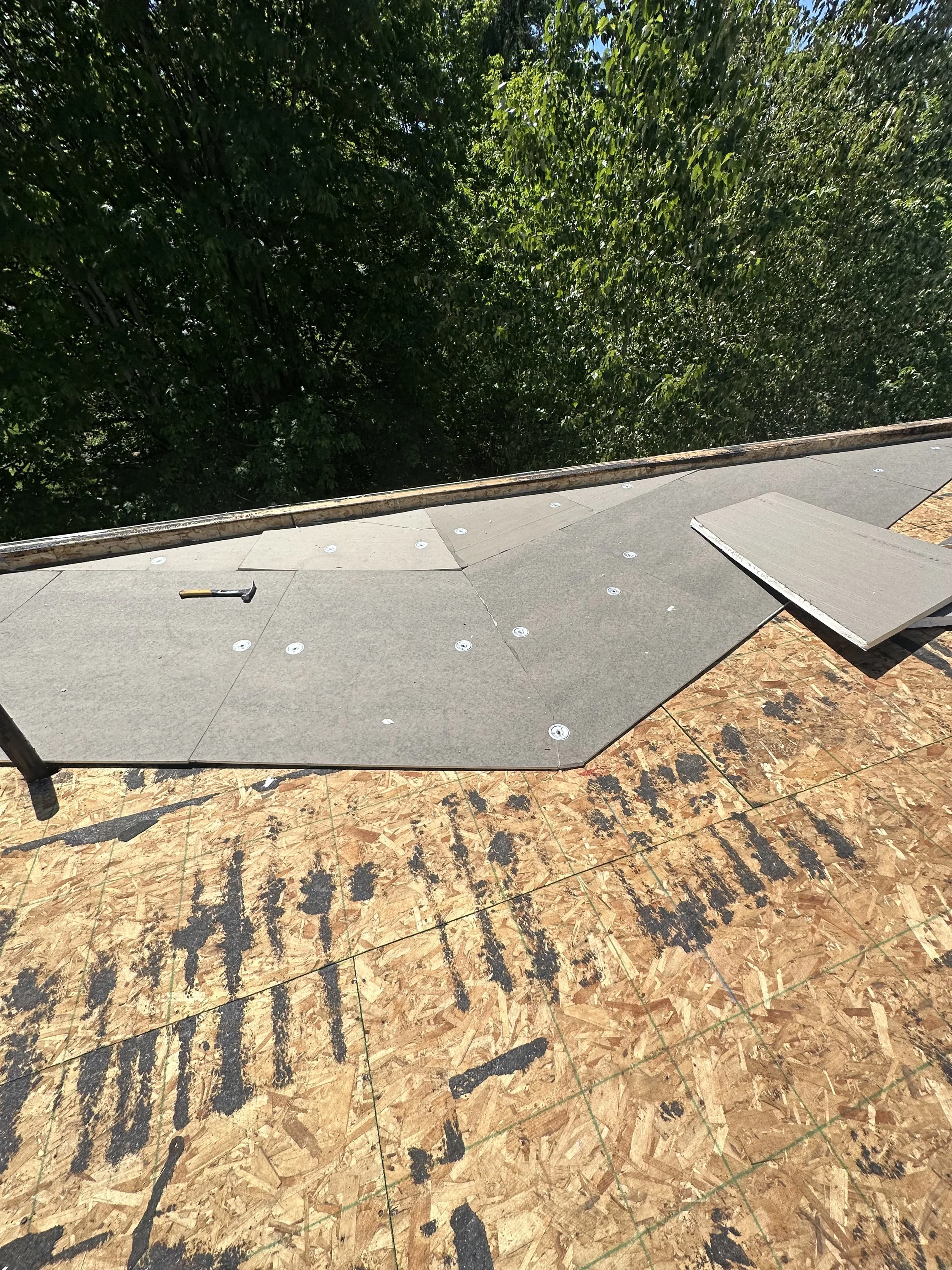 Roof under construction with installed shingles, a hammer on the roof, and plywood sheathing visible underneath.