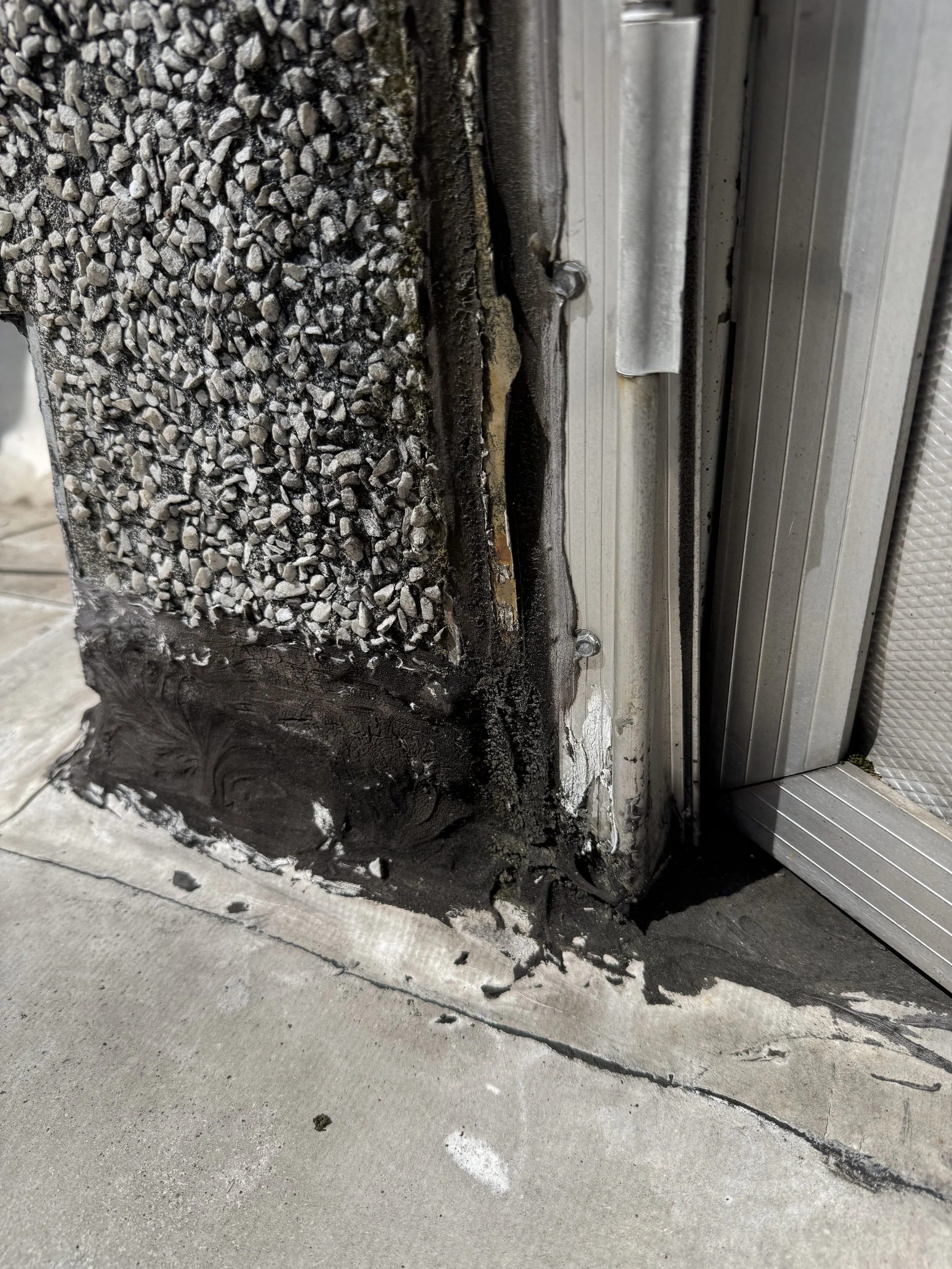 Close-up of a door threshold showing damaged insulation and wet sealant around the bottom edge of a metal door frame, with some gravel and concrete visible nearby.