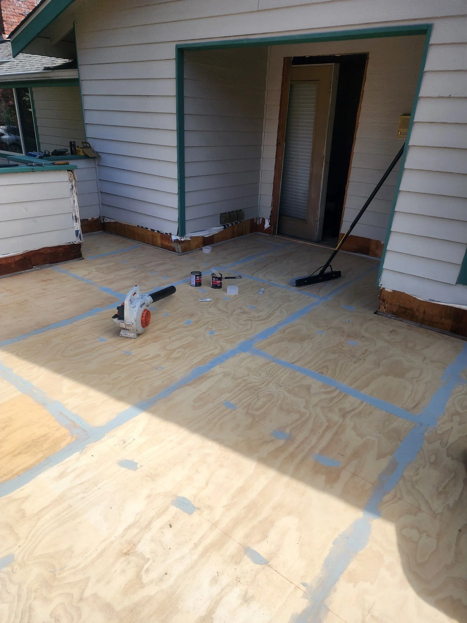 A patio or deck area under construction with plywood flooring, a power saw, cans of paint, and a broom, with the exterior of a house with white siding and a sliding door leading inside.