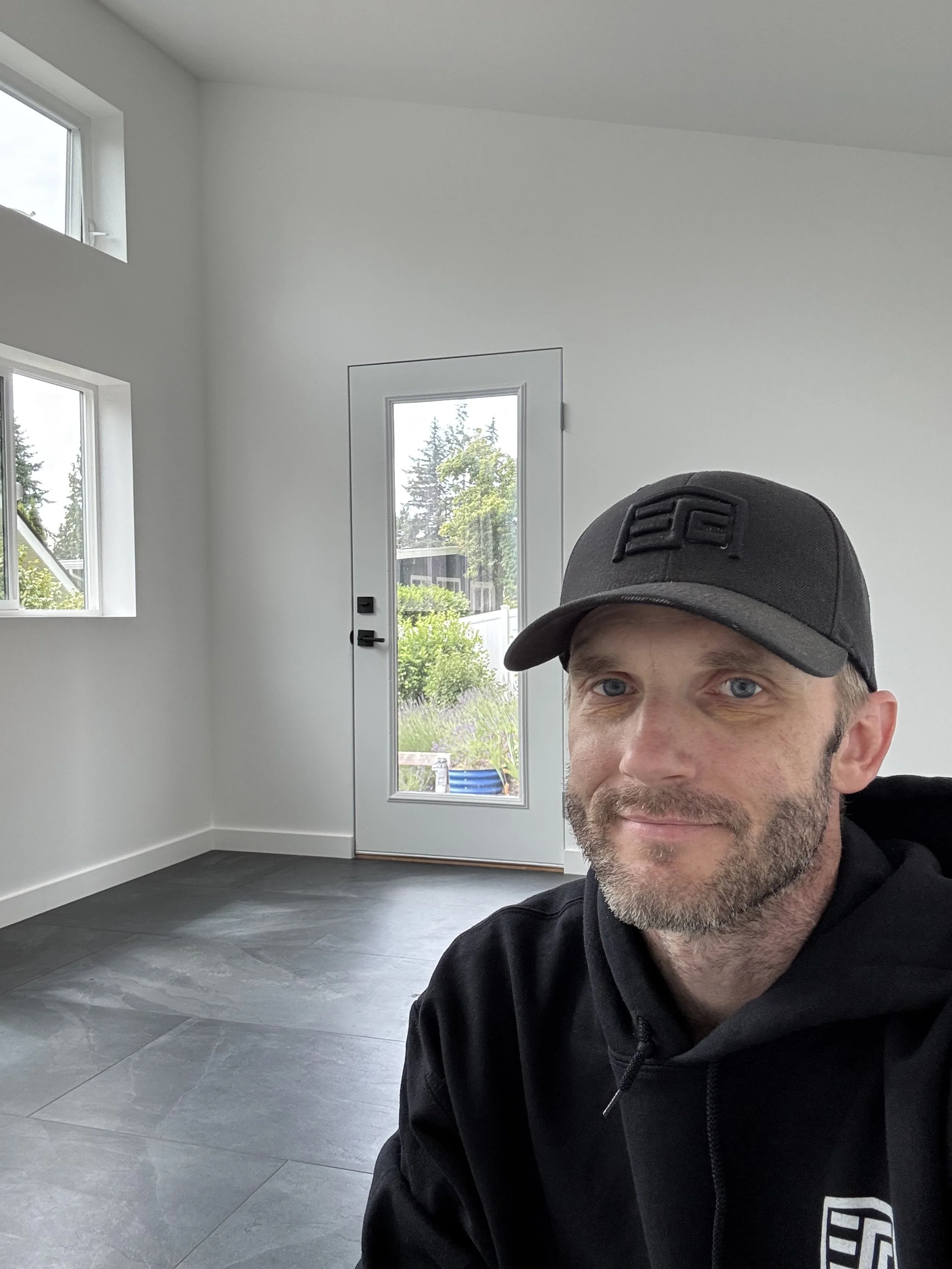 A man with a beard wearing a black hoodie and a black baseball cap taking a selfie inside a bright, empty room with white walls, large gray tile floors, windows, and a door with a window showing trees outside.