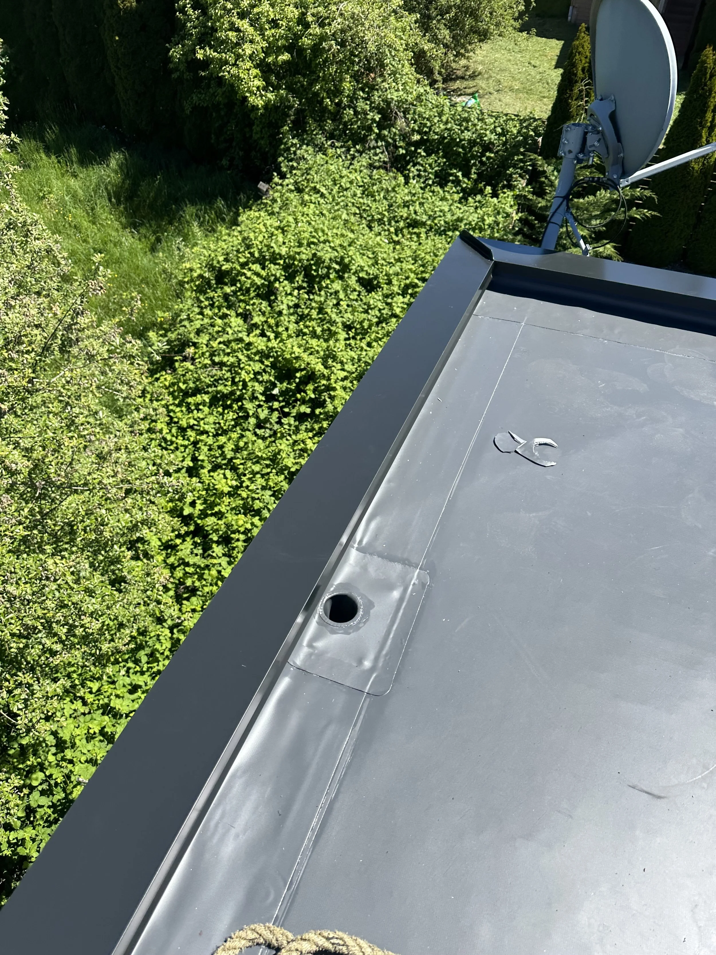 A rooftop with a satellite dish, a vent opening, and green trees in the background.