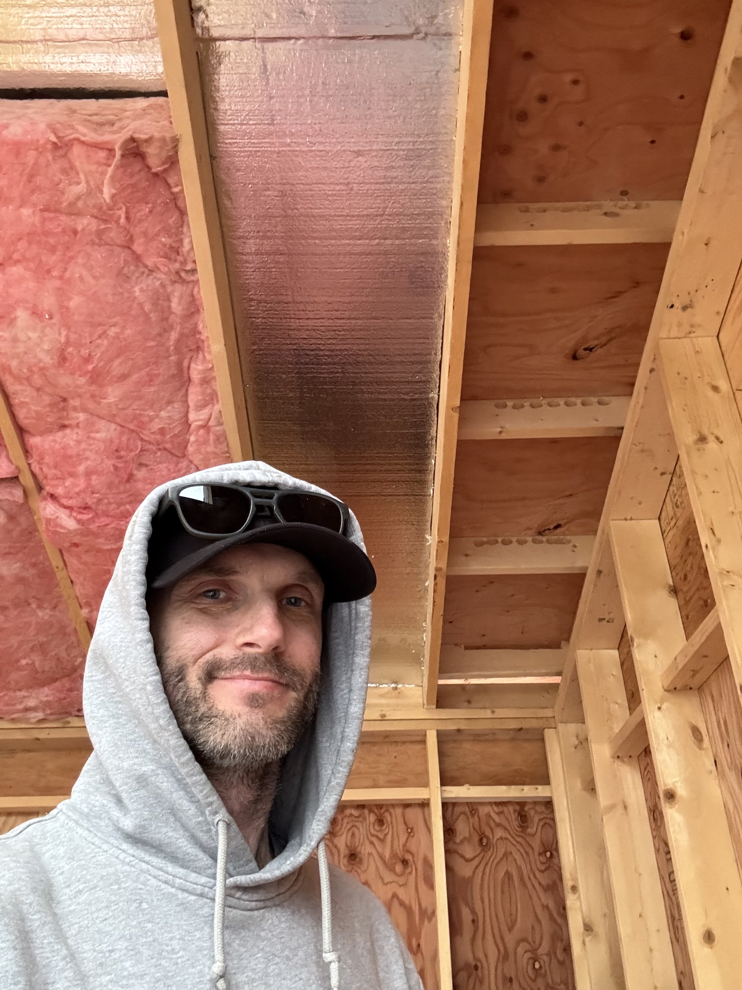 Man wearing a gray hoodie, black cap, and sunglasses on his hat, taking a selfie inside a wooden framed room under construction.