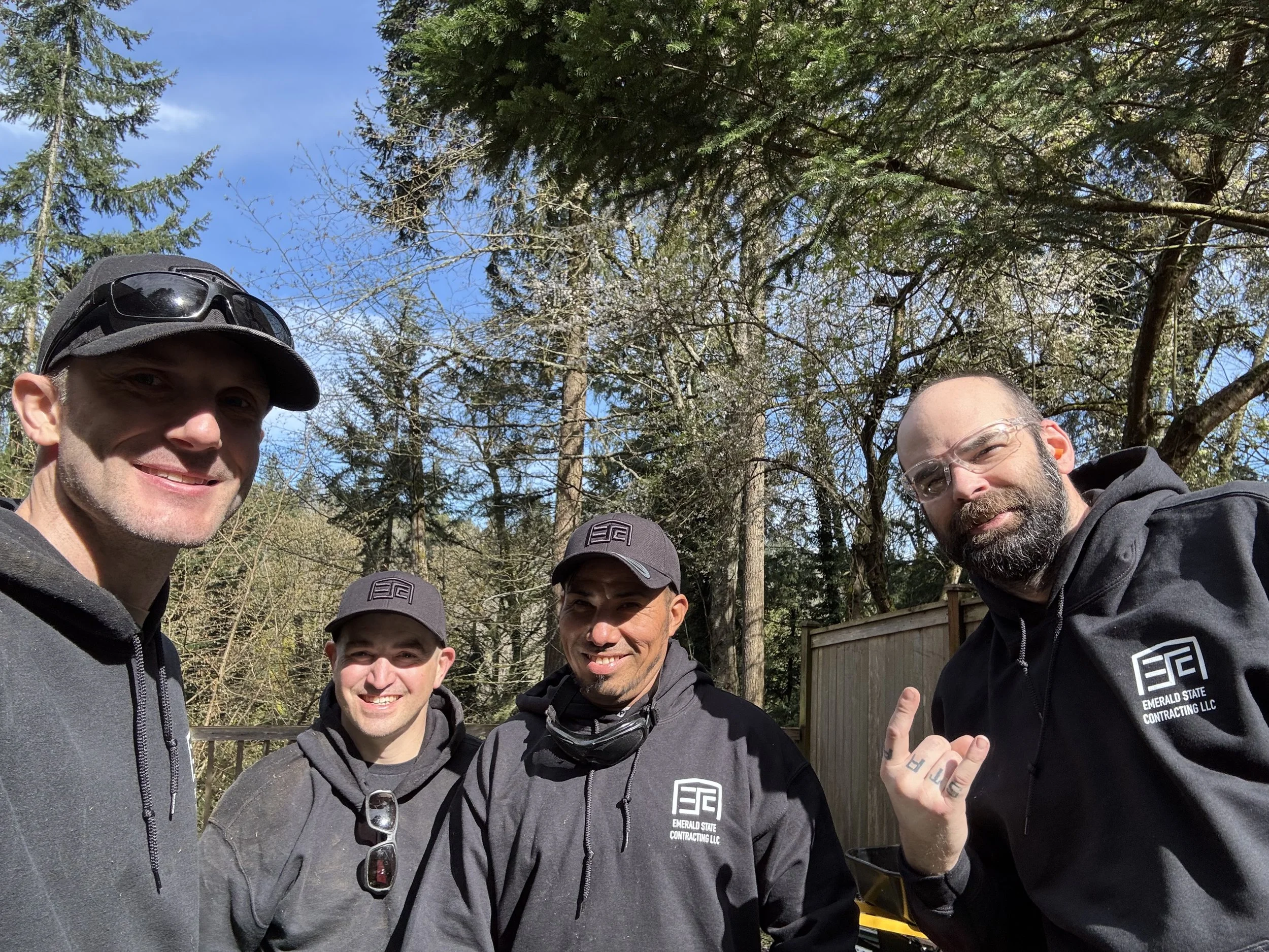 Four men smiling outdoors on a sunny day, wearing dark hoodies with 'Emerald State Contracting LLC' logo, surrounded by trees.
