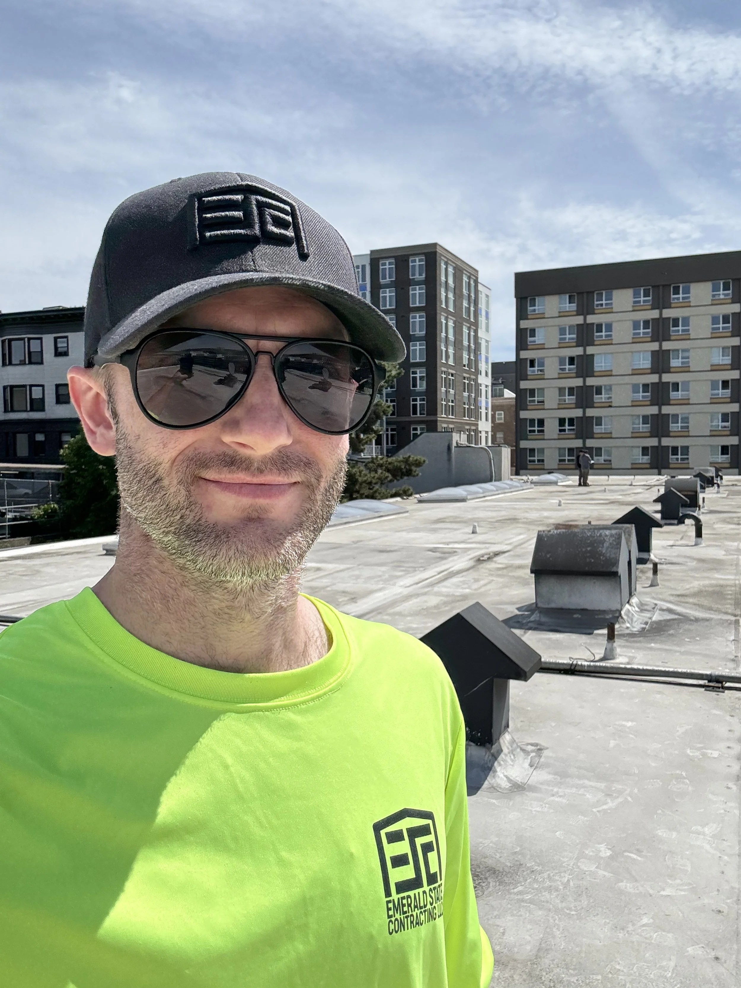 Man wearing black sunglasses, a black and gray cap, and a bright yellow T-shirt with the logo and name 'Emerald State Contracting LLC' on the front, standing on a rooftop with city buildings in the background.