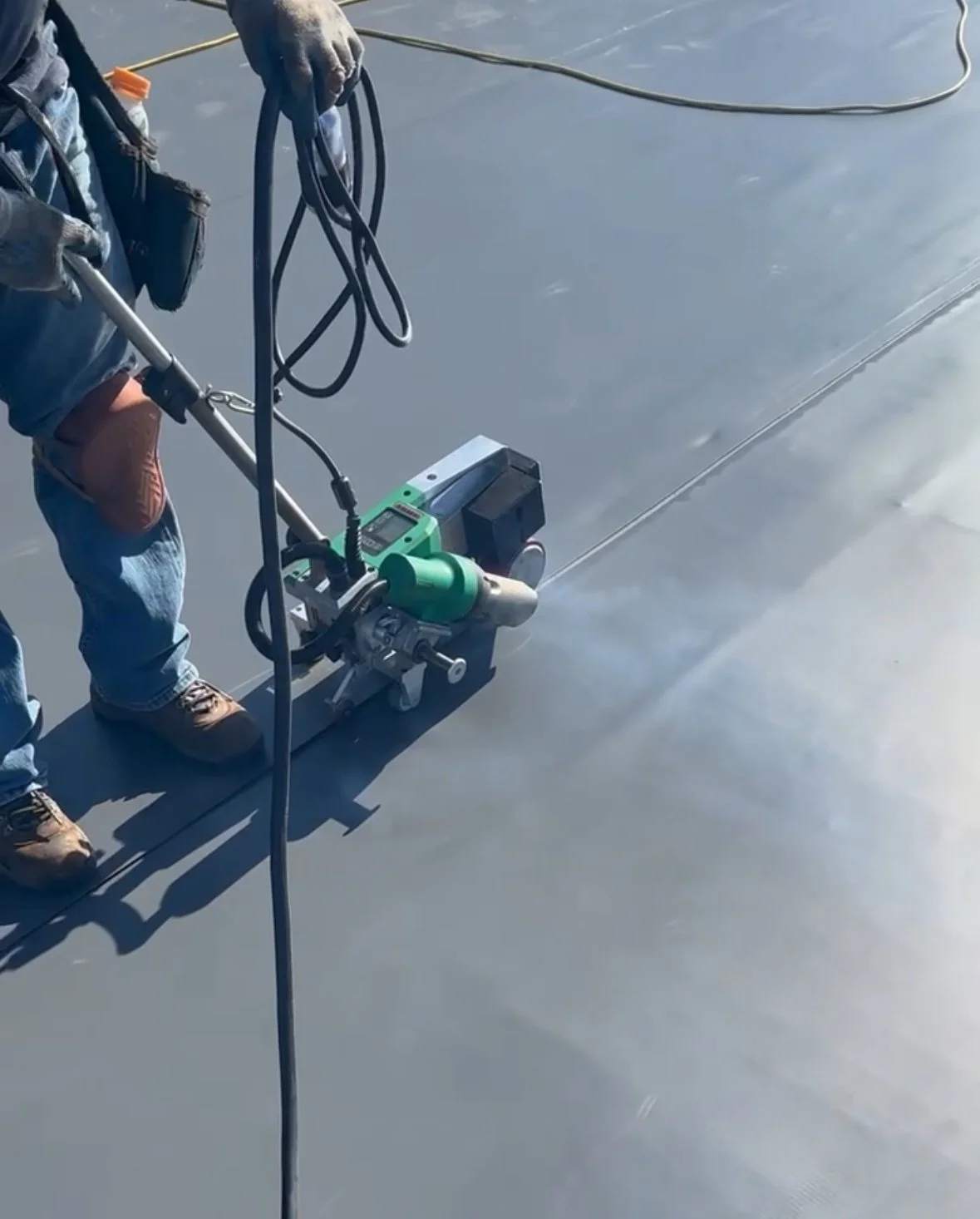 Worker using a machine to apply a smooth, gray epoxy coating on a floor, with construction tools and cords nearby.
