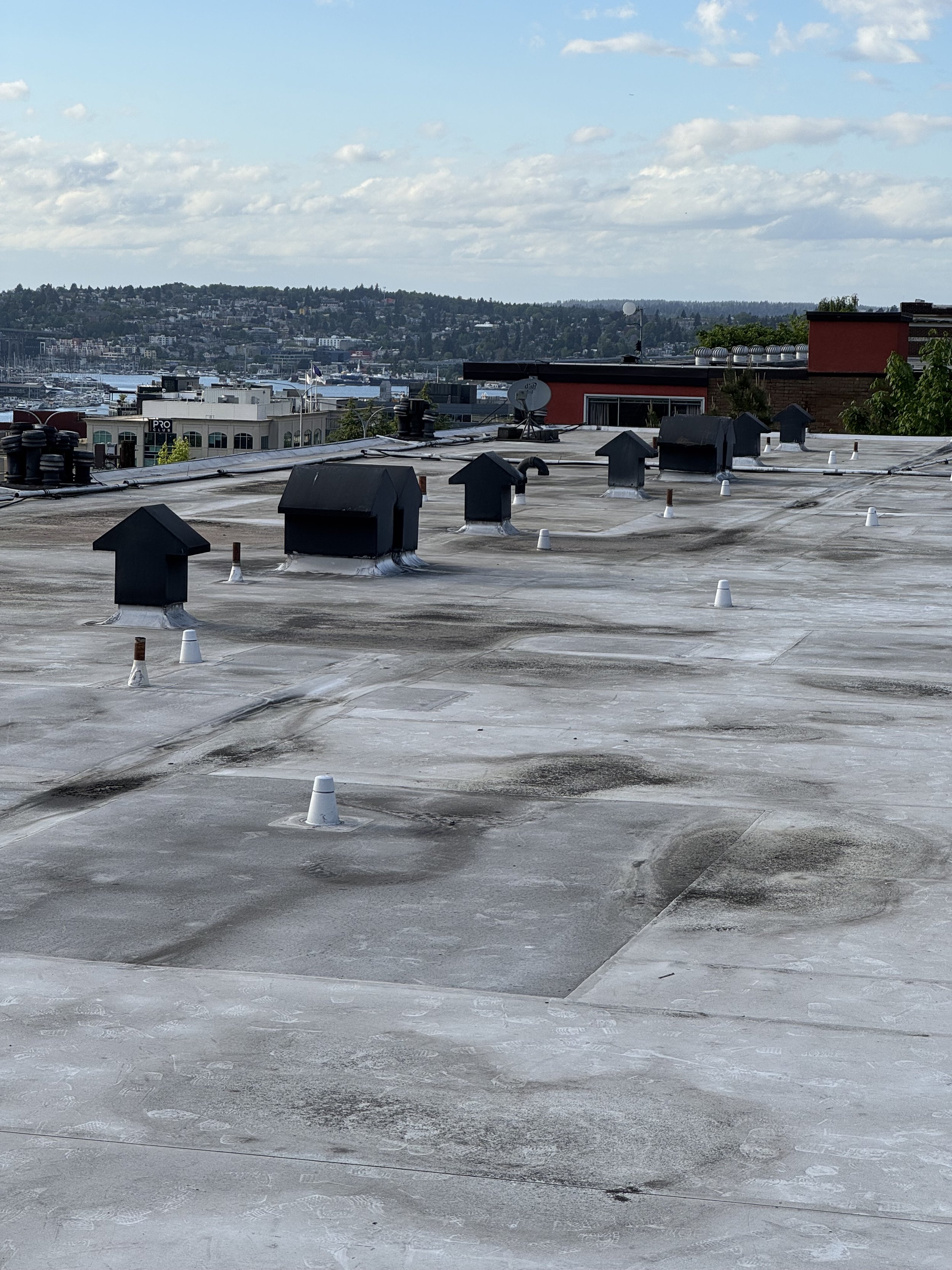 View of a flat rooftop with multiple black vent caps, white cones, and patches of wear and discoloration. In the background, there are buildings, a body of water, and hills under a partly cloudy sky.