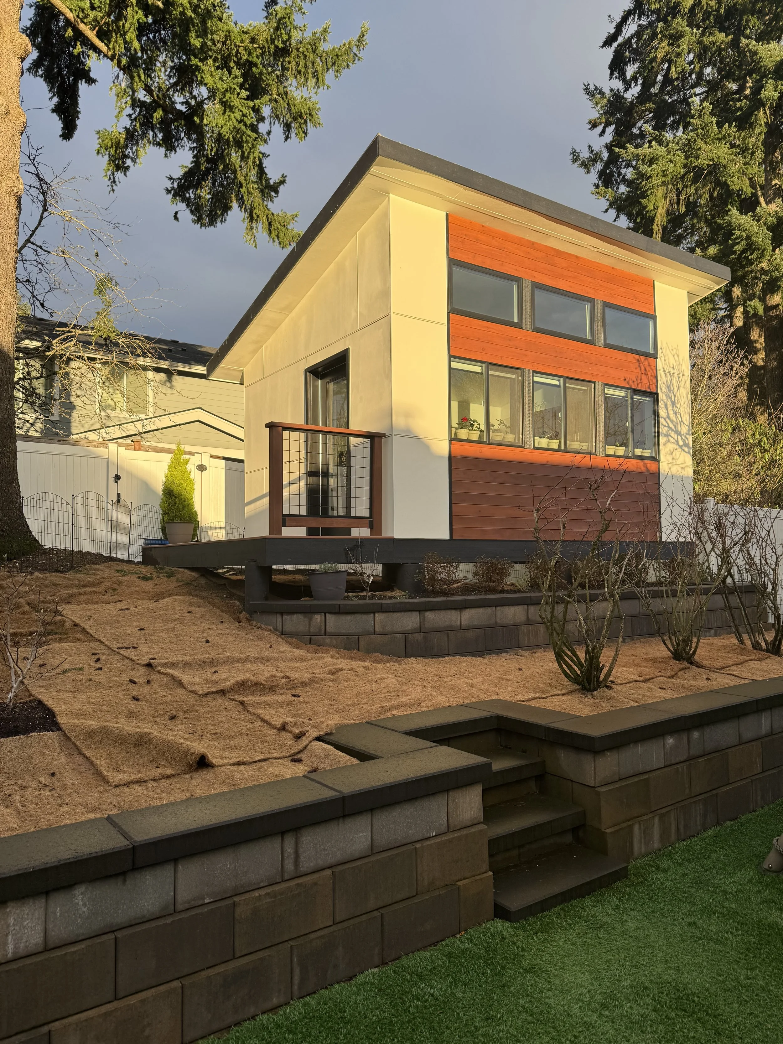 Modern two-story house with large windows, white and wooden exterior, and a small balcony, surrounded by landscaped yard with trees and shrubs.