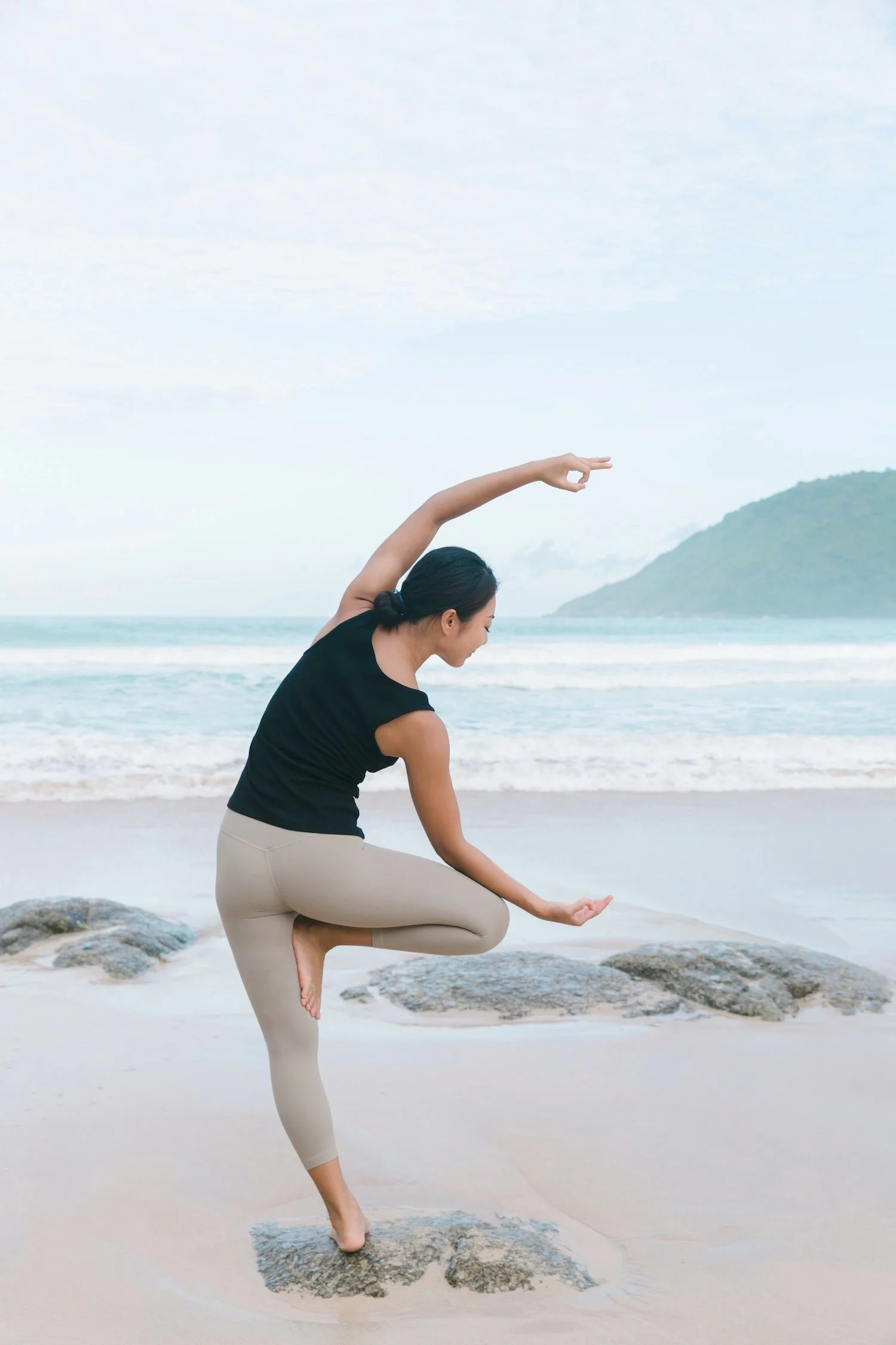 A woman practicing yoga on the beach, balancing on one foot with her other foot resting on her inner thigh, and arms in a circular position, near rocks and the ocean with a mountain in the background.