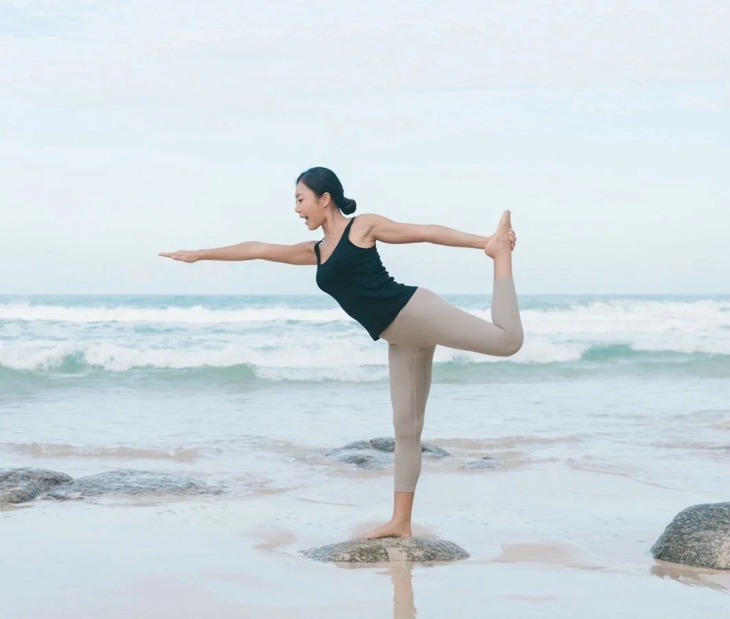 Woman practicing yoga on the beach in a standing balancing pose, with ocean waves in the background.