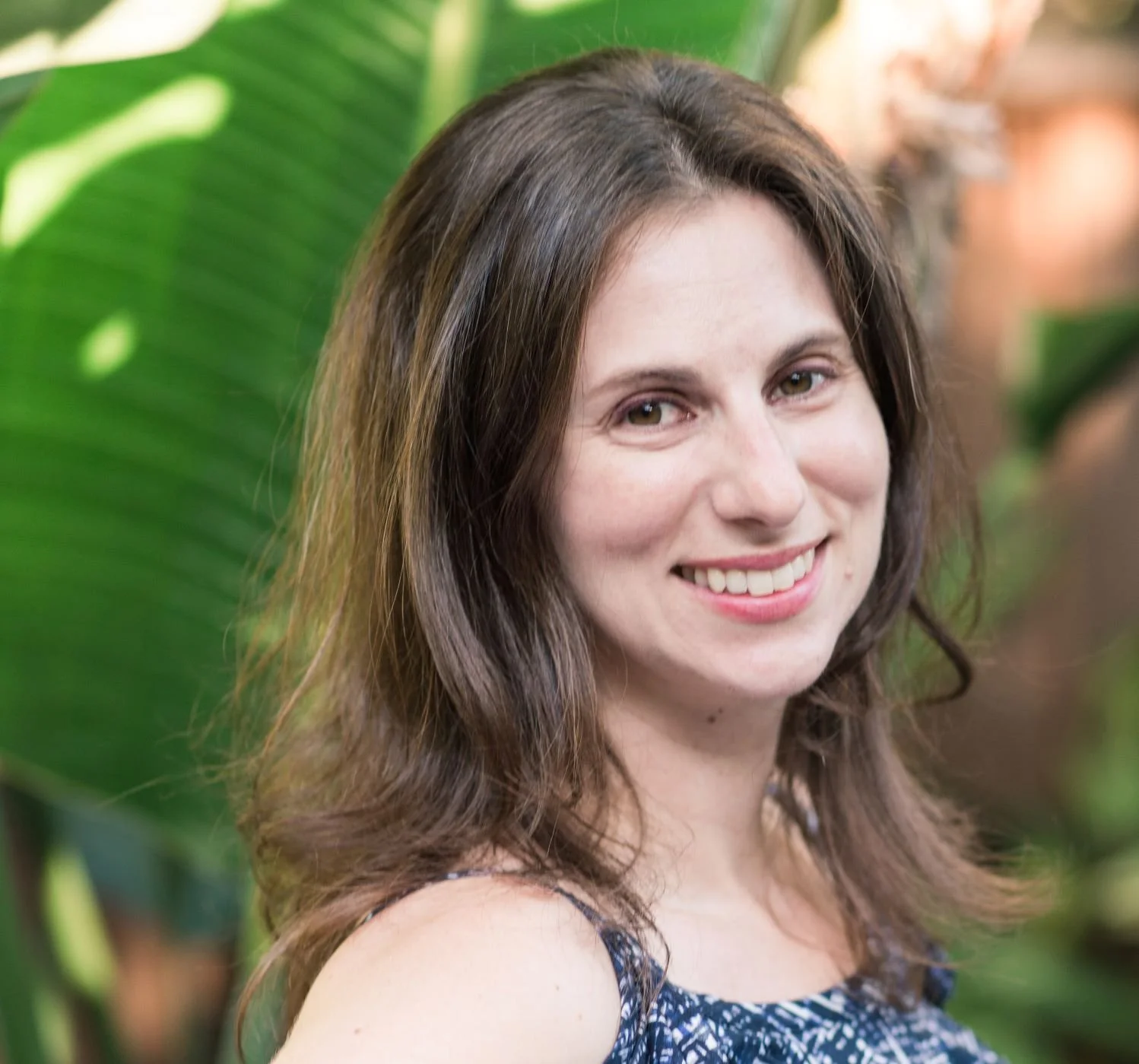 Smiling woman with brown, wavy hair standing outdoors near large green tropical leaves.
