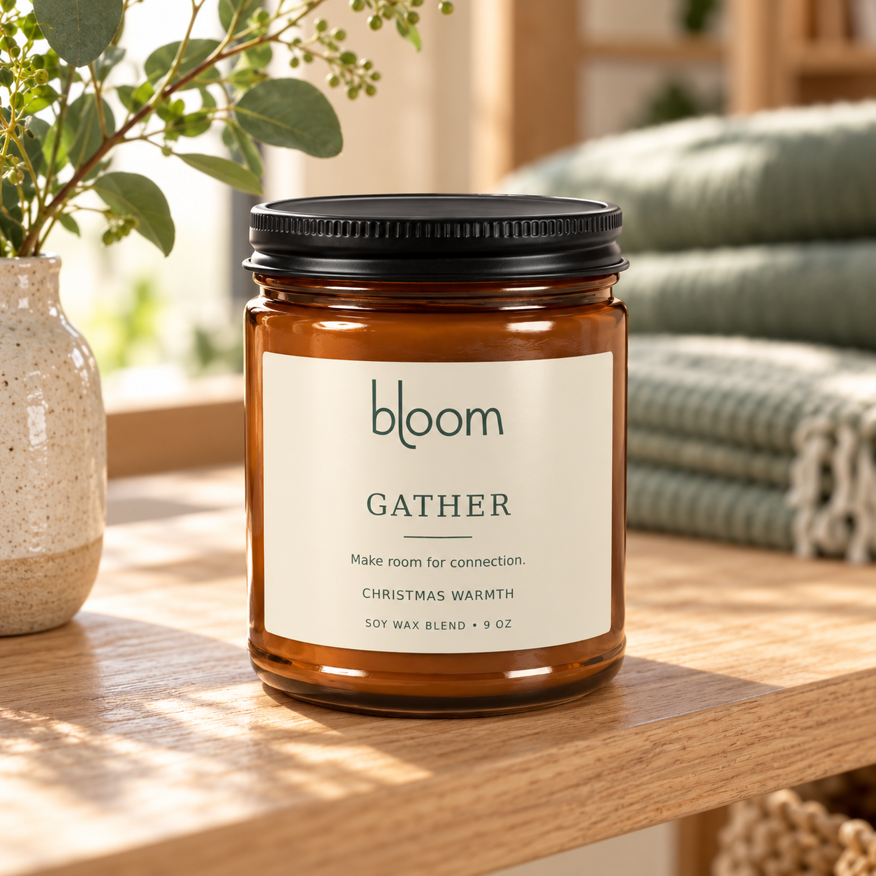 Bloom Gather 9 oz Christmas Warmth candle styled on a table beside a vase and folded blanket.