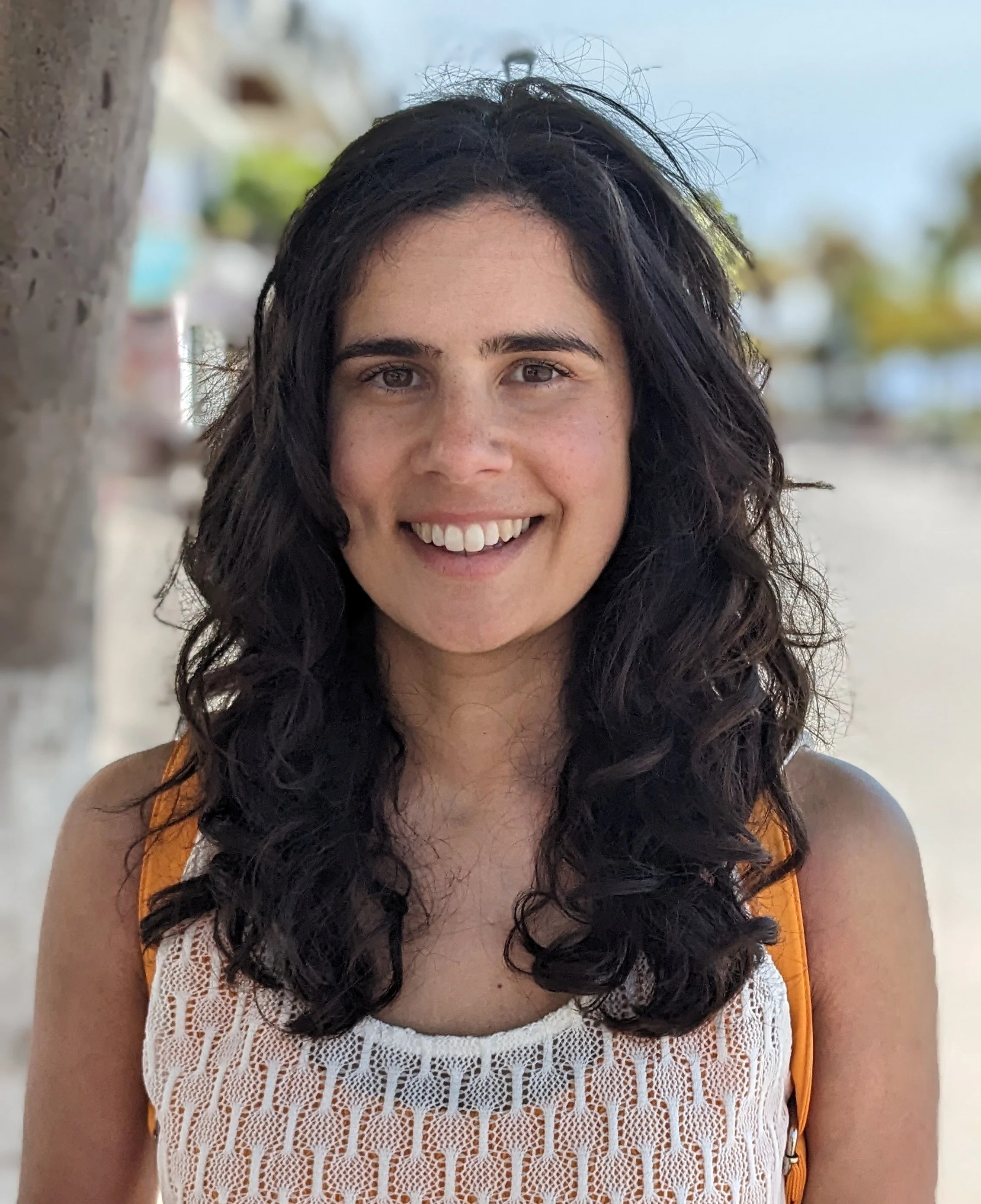 A smiling woman with dark, curly hair wearing a white crocheted top and an orange sleeveless jacket, standing outdoors near a tree with a blurred background of trees and sky.