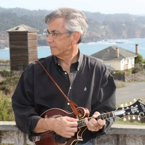 A man with gray hair and glasses playing a mandolin outdoors near the coast, with houses and hills in the background.