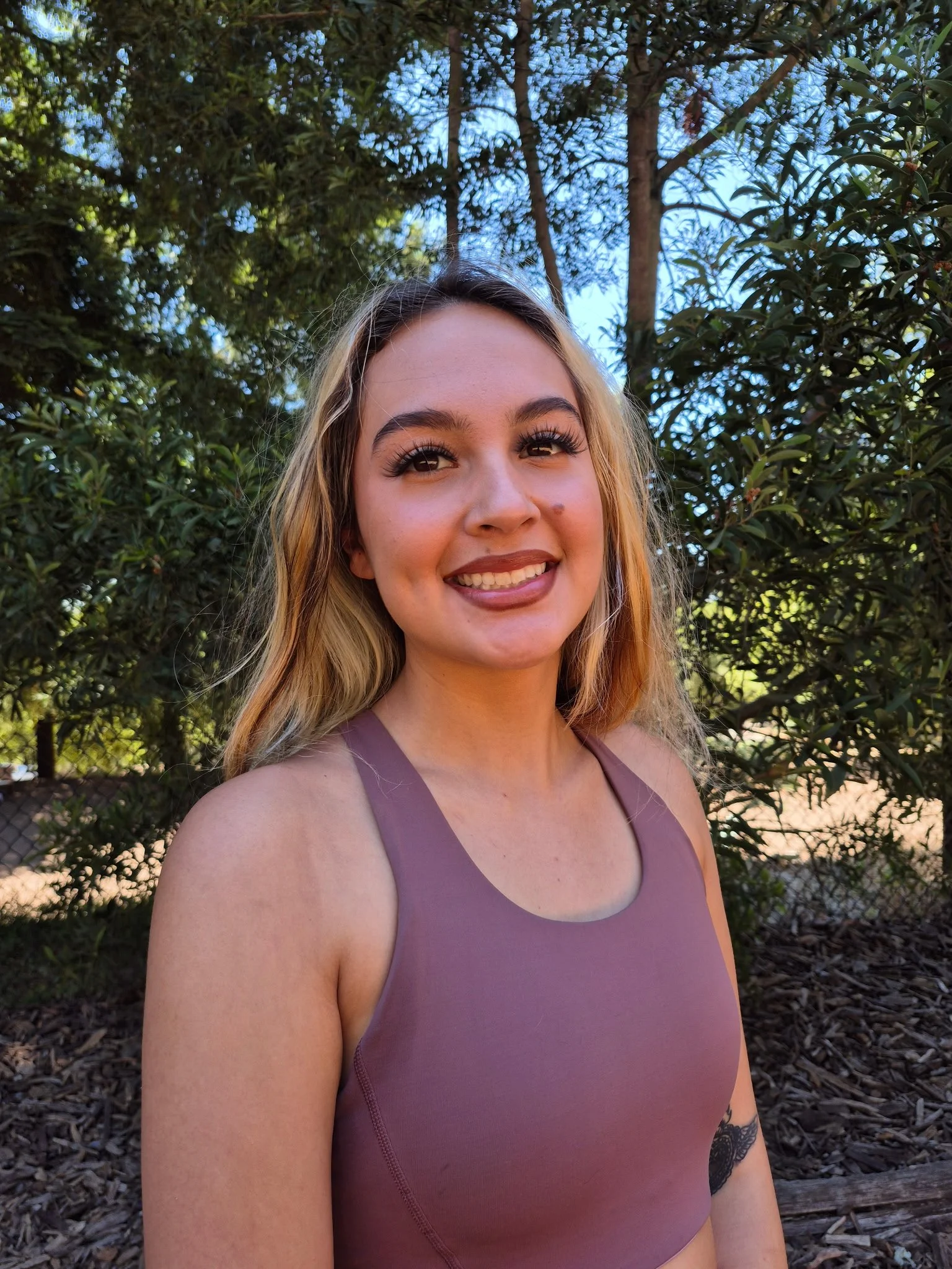 A woman with long blonde hair, smiling, standing outdoors in front of trees and foliage, wearing a mauve sleeveless top.