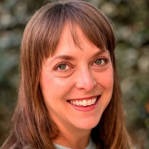 Close-up of a woman with brown hair, smiling outdoors.