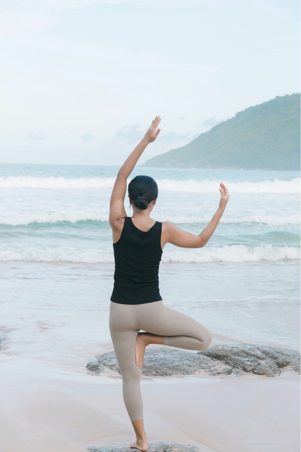 Person practicing yoga on the beach near the ocean, performing a tree pose with arms raised and one foot resting on the inner thigh.