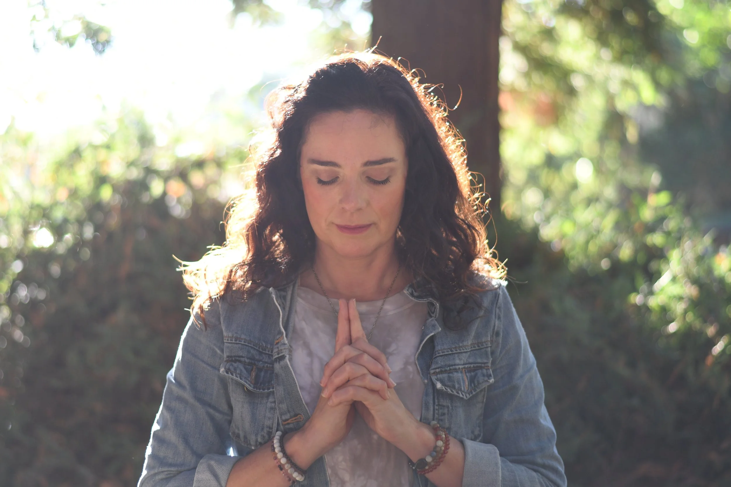 A woman with curly dark hair that is backlit by sunlight, standing outdoors with her eyes closed and hands pressed together in a prayer position.