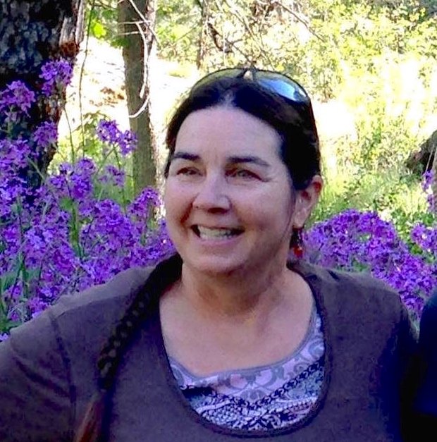 Smiling woman with dark hair in braids outdoors, surrounded by purple flowers and greenery.