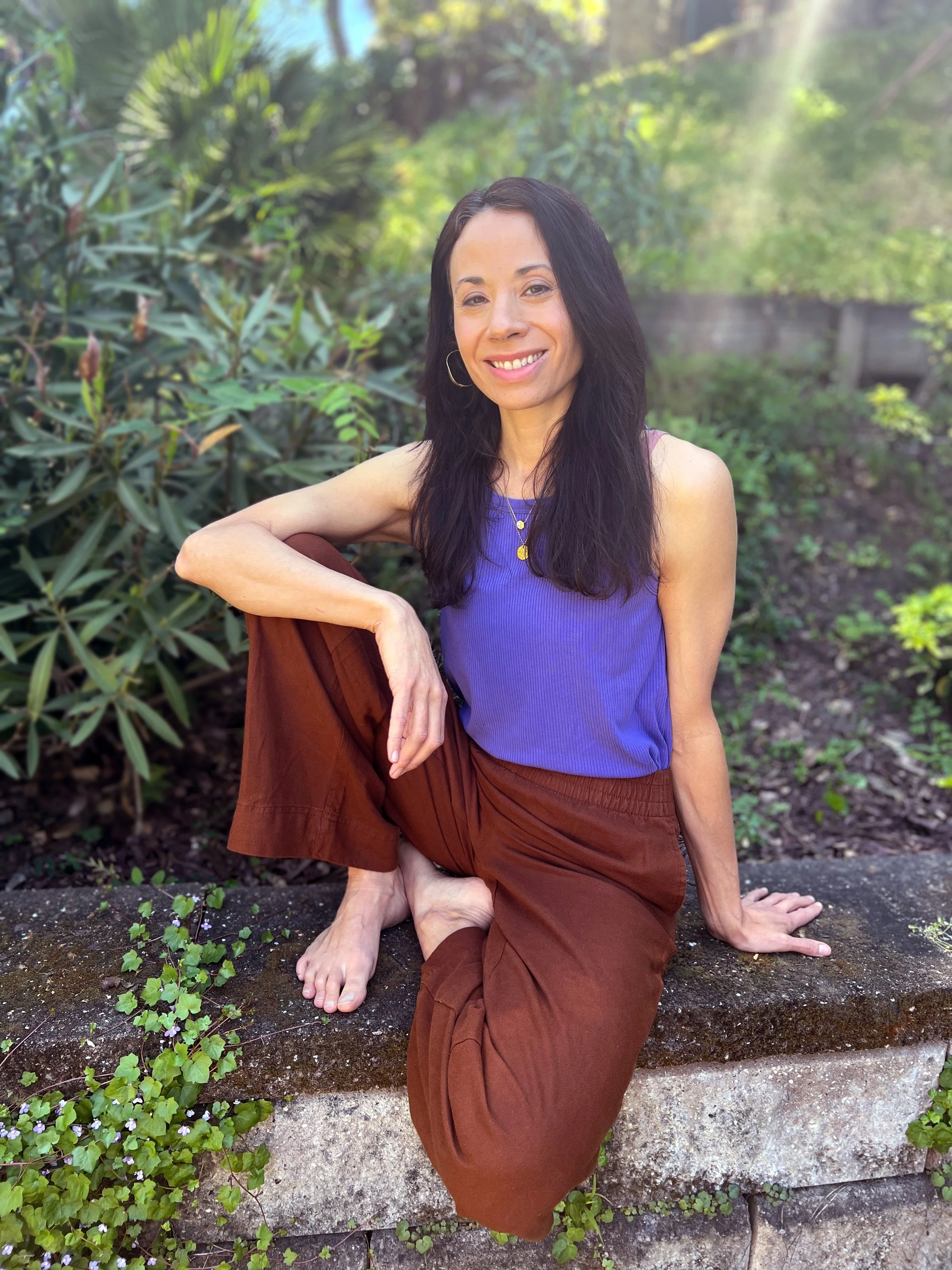 A woman with long black hair, wearing a purple sleeveless top, gold jewelry, and loose brown pants, sitting outdoors on a stone ledge with one leg crossed and barefoot, surrounded by greenery and sunlight.