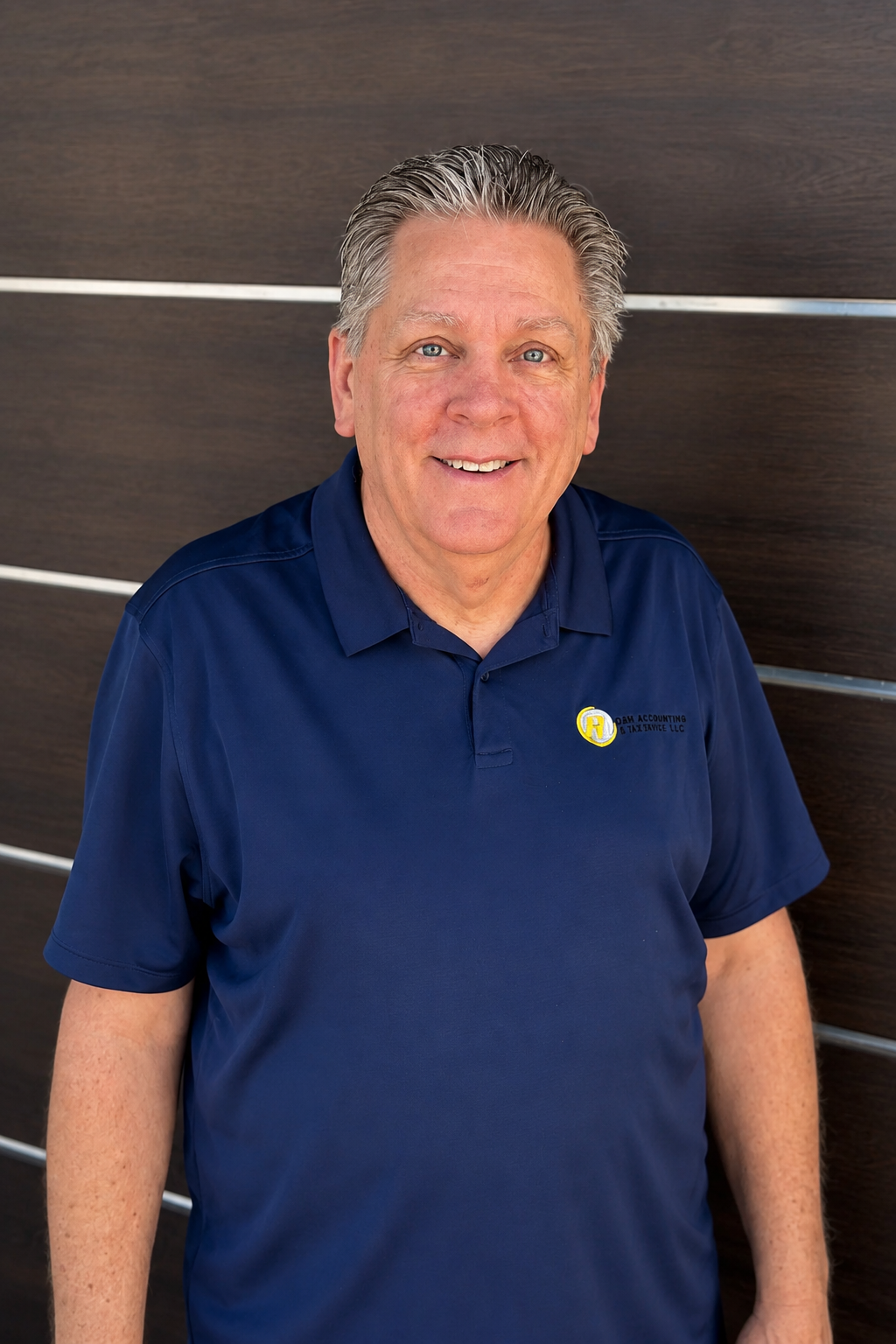 A smiling older man with gray hair and blue eyes wearing a navy blue polo shirt with a company logo standing against a dark wood-paneled wall.