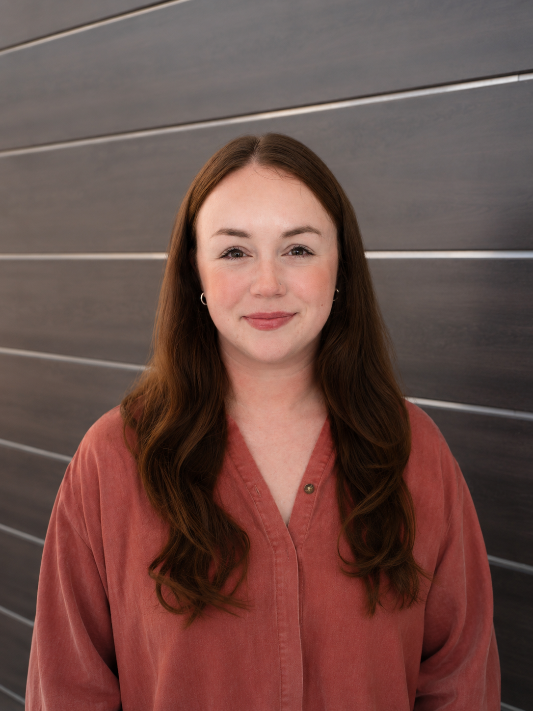 A woman with long, wavy brown hair wearing a reddish-brown button-up shirt, smiling at the camera, standing against a dark horizontal wooden wall.