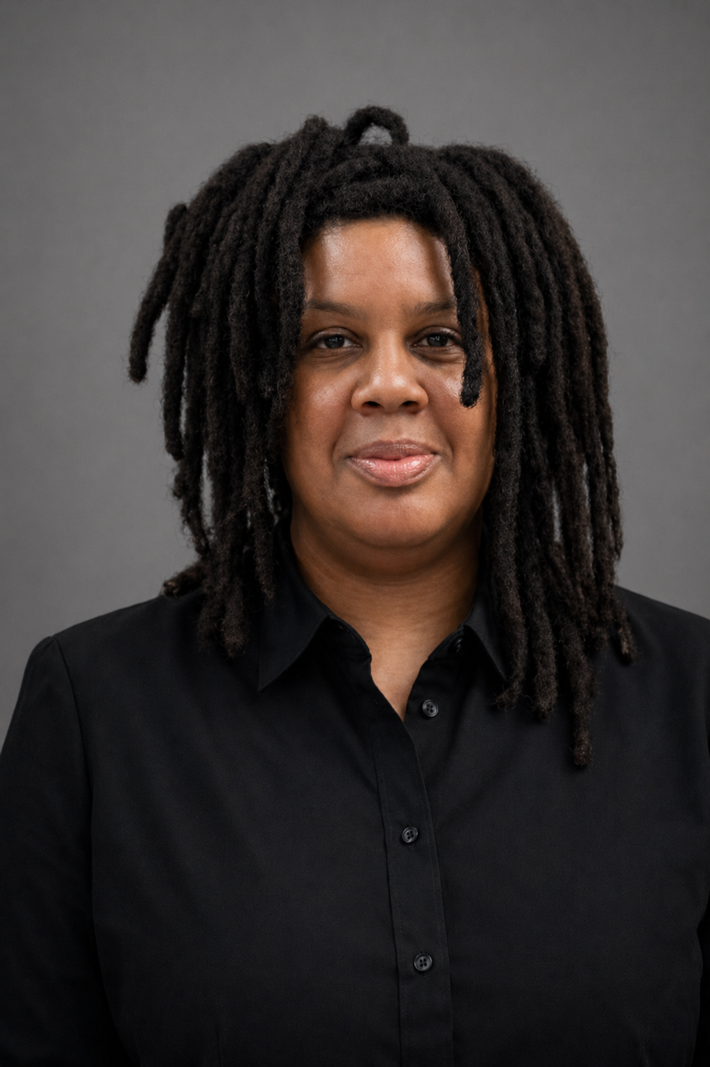 Persona con cabello de rastas, camisa negra y collar, frente a una pared de ladrillos grises.