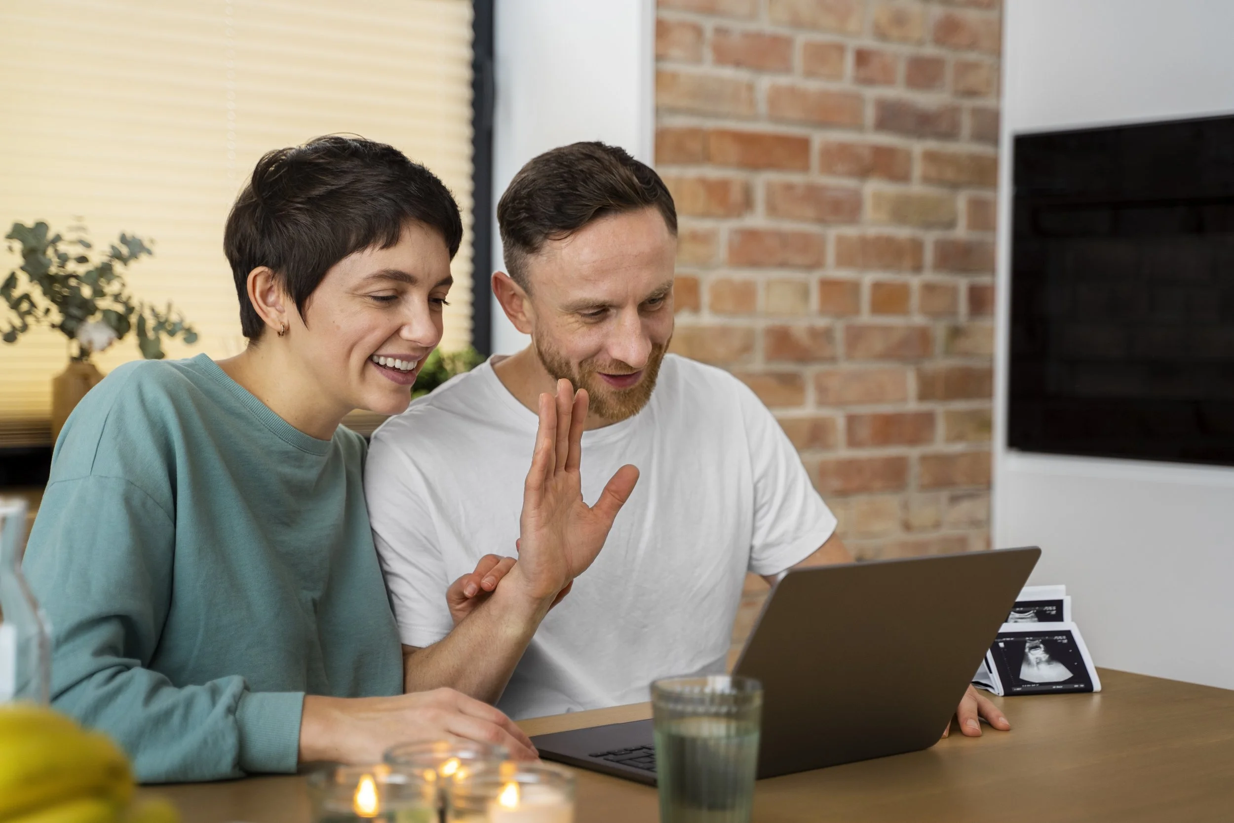 Pareja sonriendo mientras usan una computadora portátil, con ecografías en la mesa.