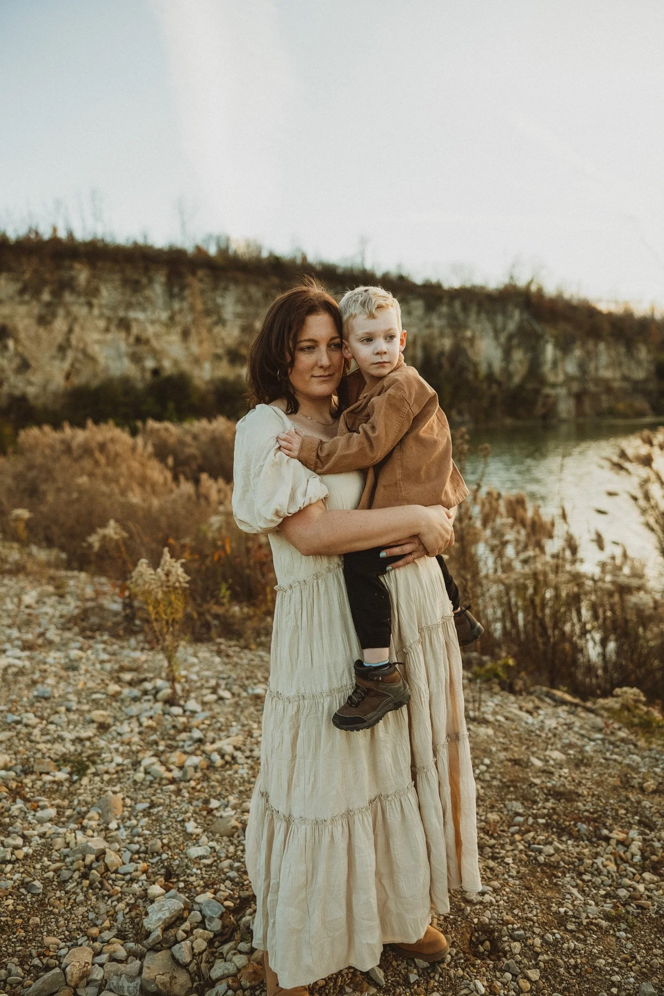 A woman holding a young boy outdoors near a river with a rocky, autumn landscape background.