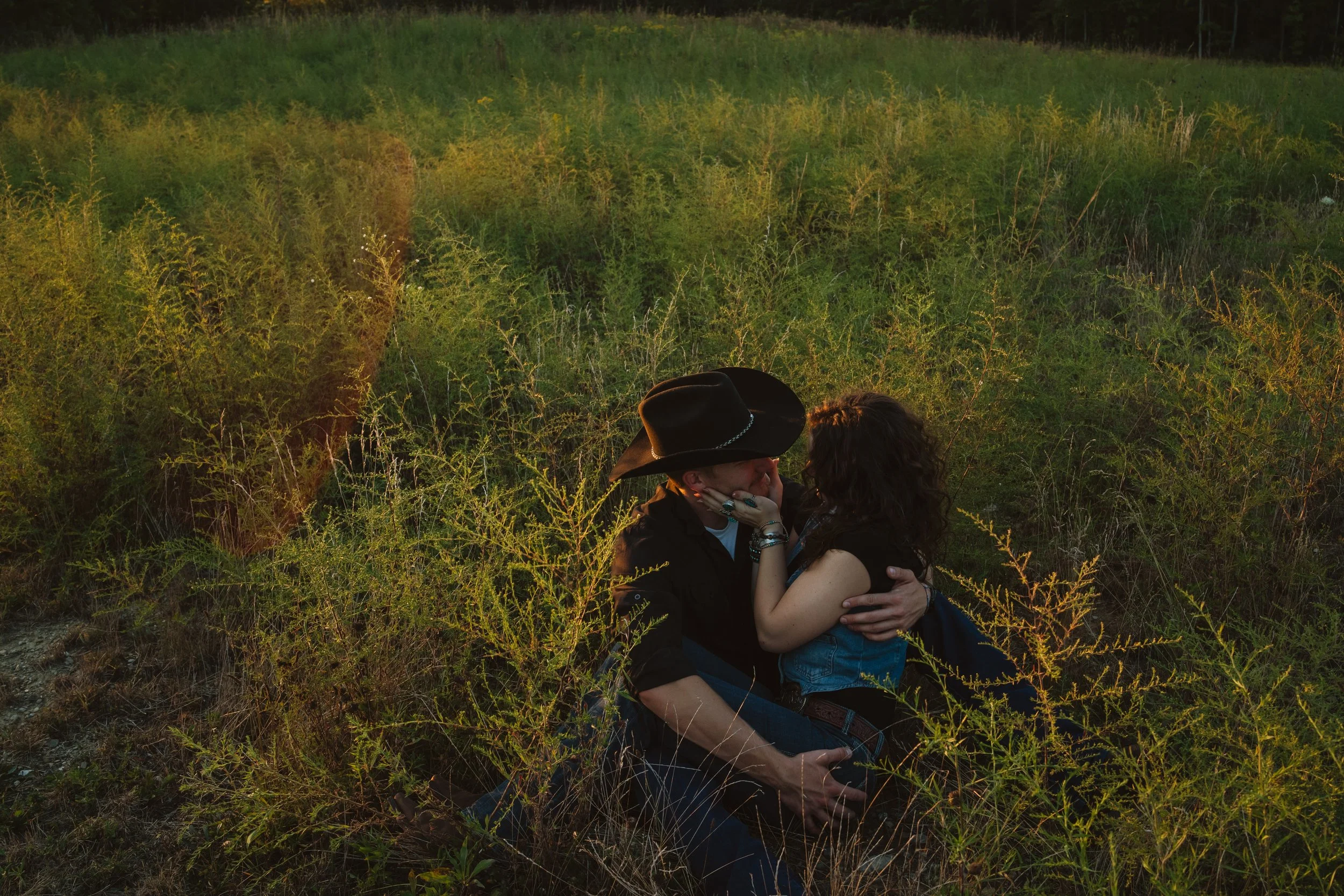 A man and woman sitting together in a grassy field during sunset, facing each other closely, with the woman touching the man's face and both showing affection.