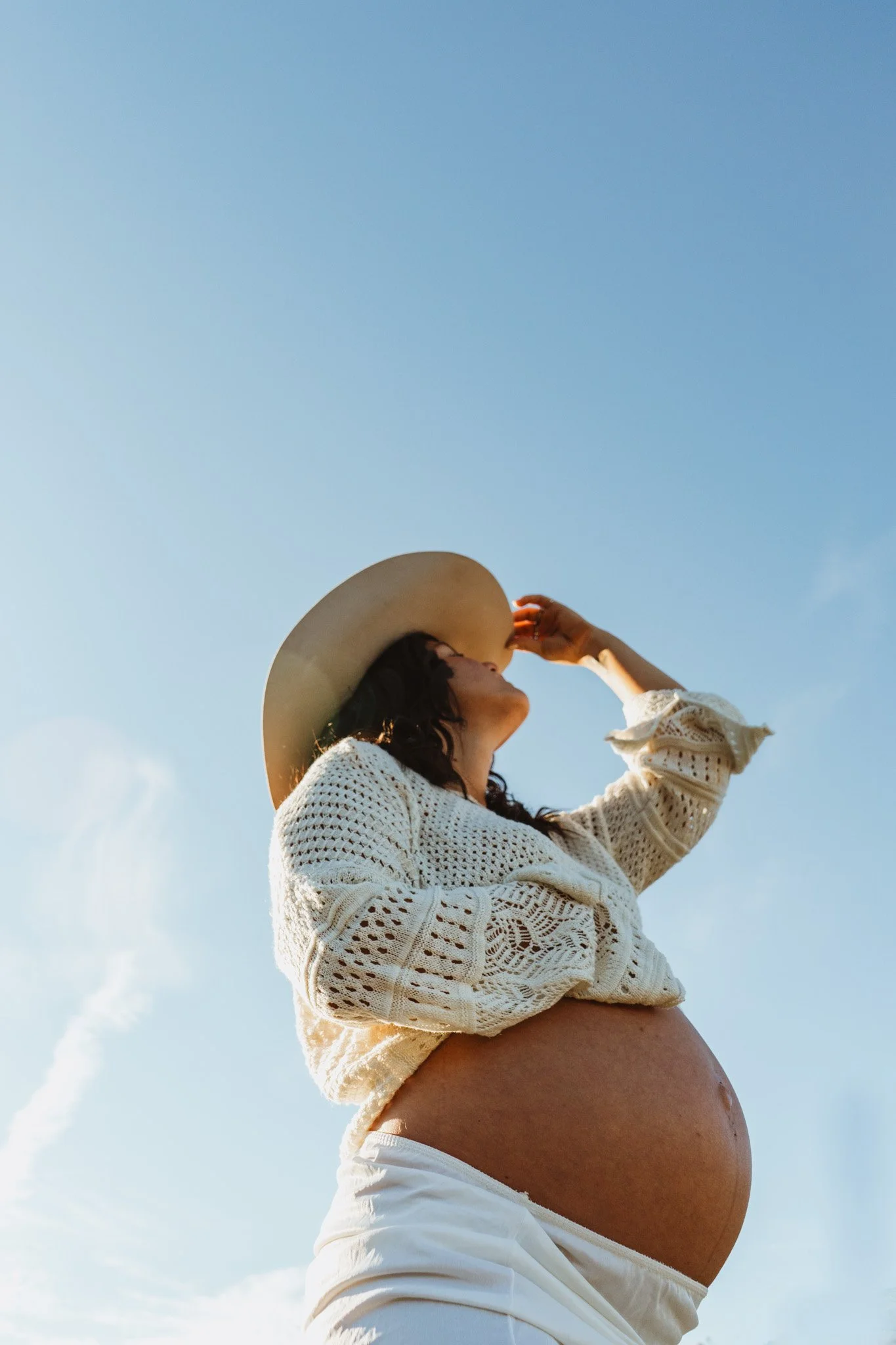 A pregnant woman in a white knitted sweater and white pants, wearing a large wide-brimmed hat, standing outdoors against a blue sky, with her hand holding the hat as she looks upward.