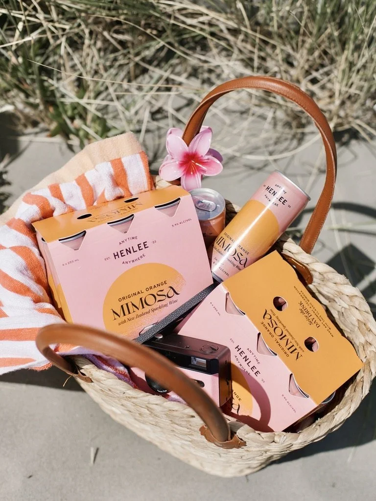A straw basket containing orange and pink canned beverages, boxes of Henlee Mimosa wine, a small bottle, a camera, a pink flower, and a striped towel, outdoors with grass and concrete visible.