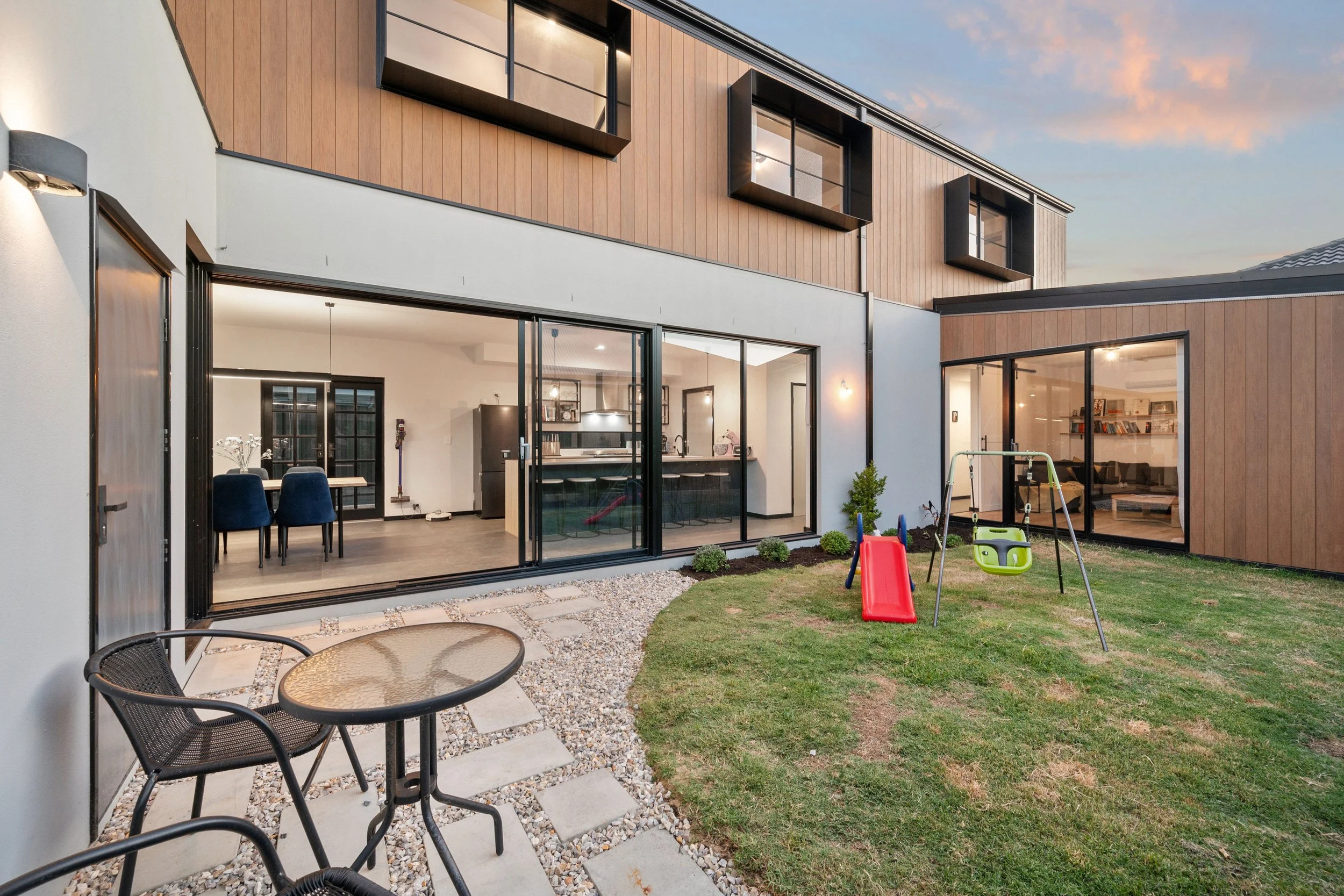 Modern sustainable high performing architectureModern house with large glass sliding doors opening to a backyard with a small swing set and a patio table with two chairs.