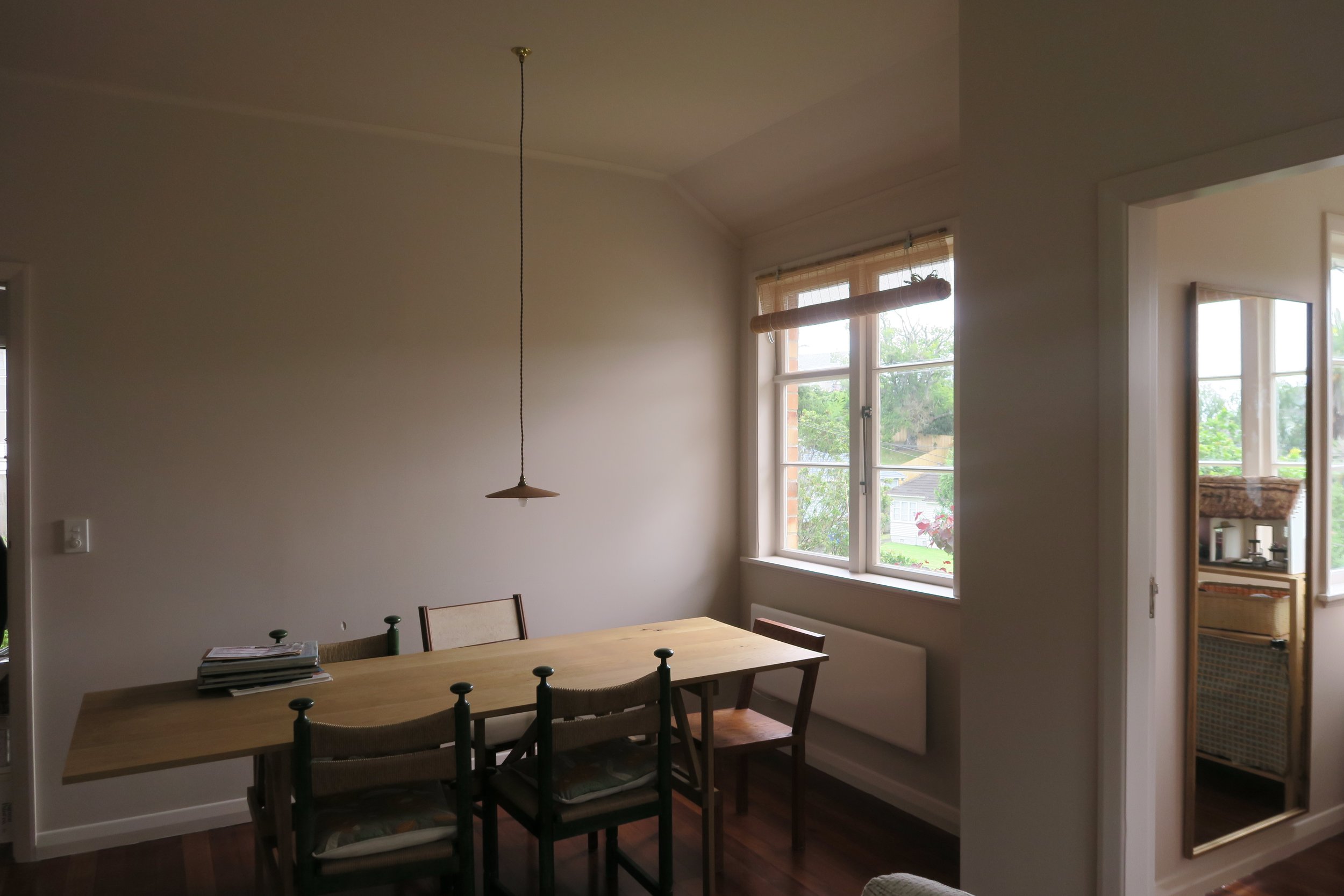 A dining room with a wooden table and four chairs, a hanging light fixture above, two windows with light-colored blinds, and a doorway leading to another room with a mirror on the wall.