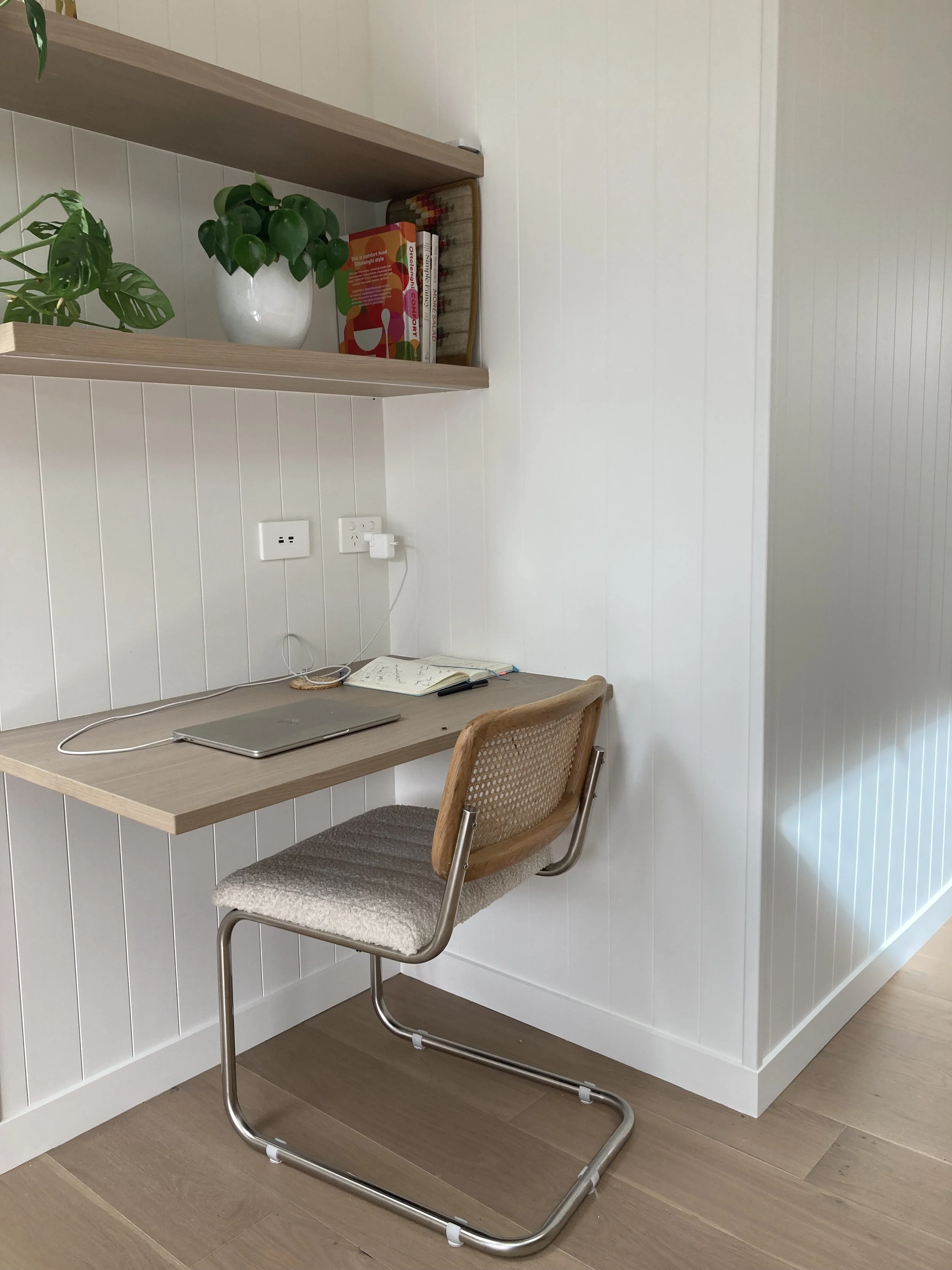 A small workspace with a wooden chair, a floating wooden desk, a closed laptop, notebook, pen, power outlet, and a plant on a shelf above the desk, all against white paneled walls and wooden flooring.