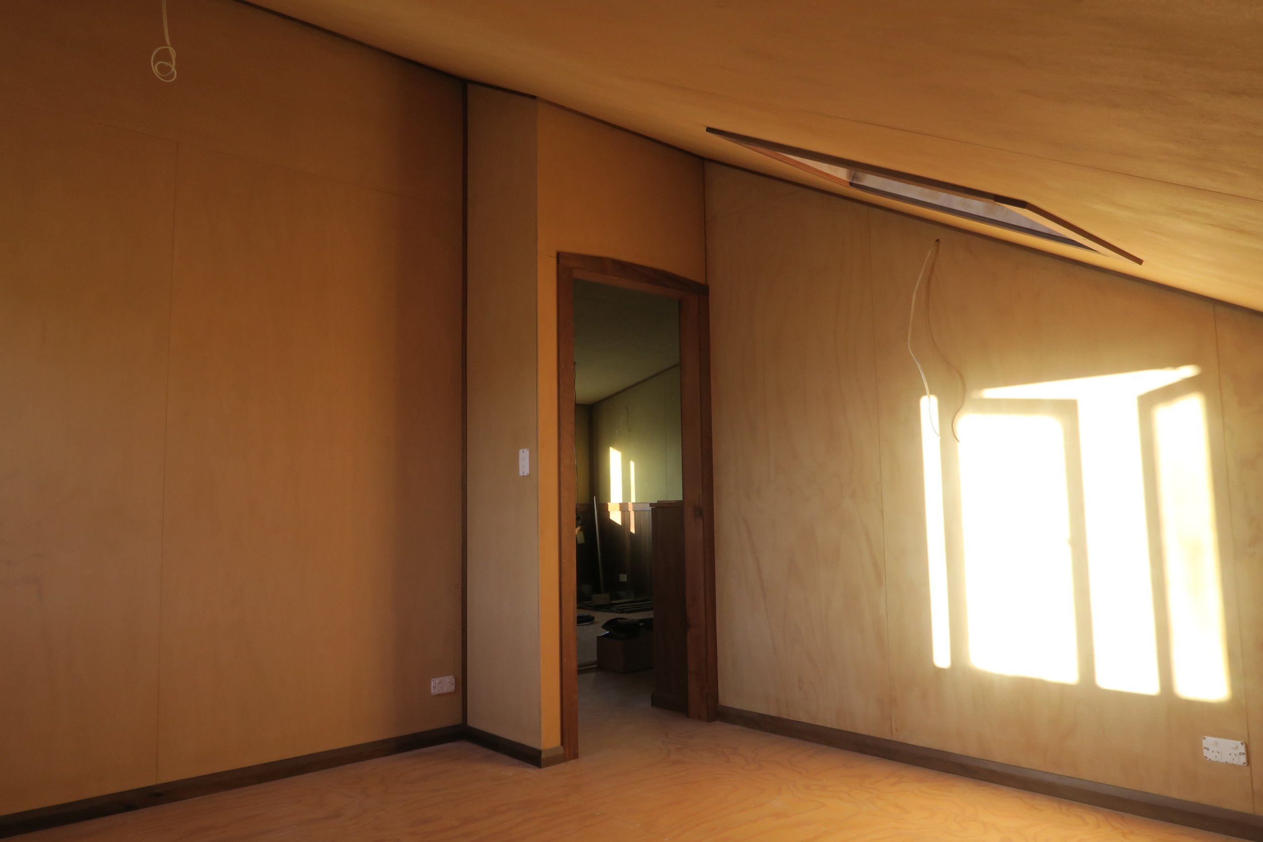 Empty room with natural sunlight, wooden walls, sloped ceiling, and an open door opening into another room.
