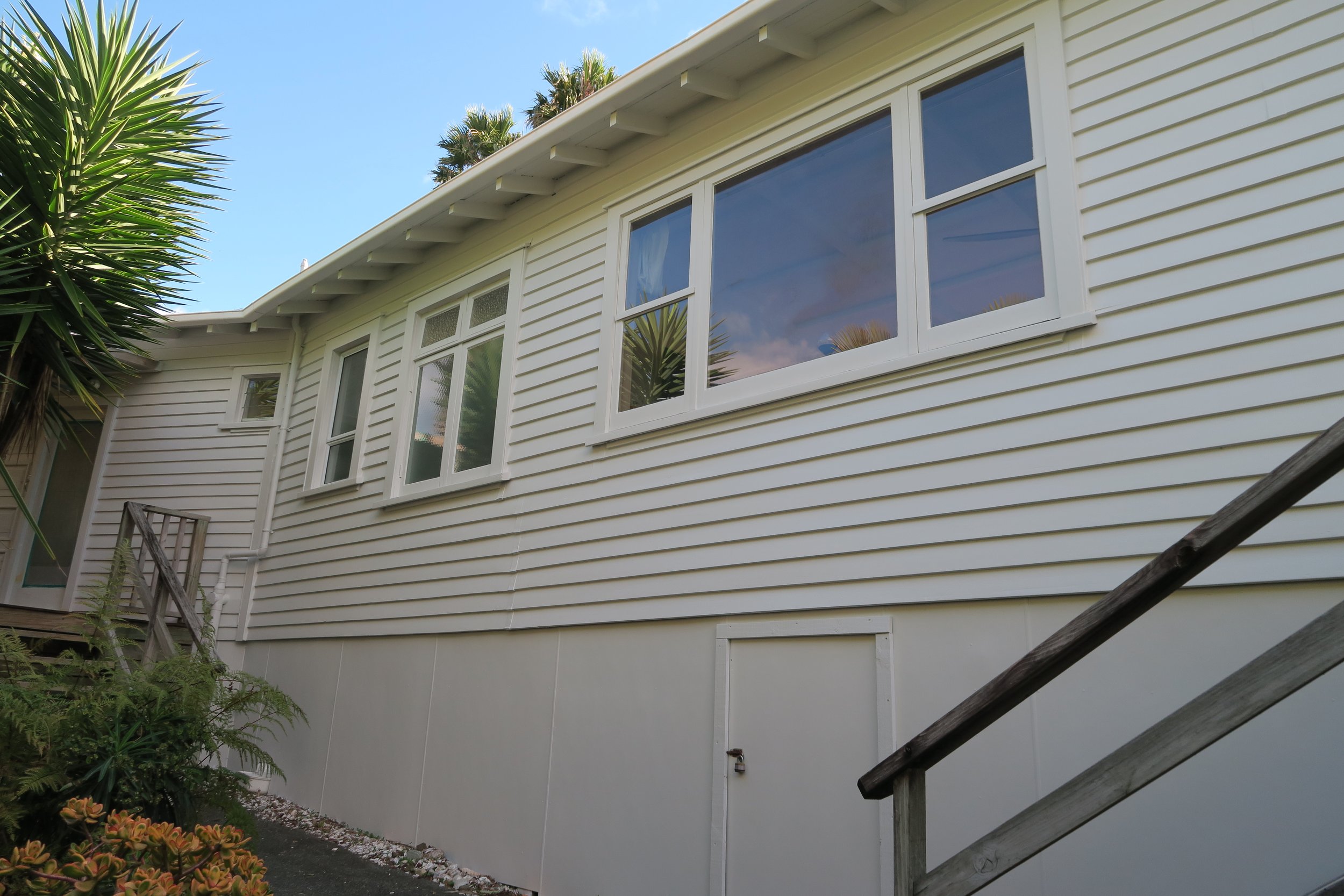 Back of a house with white siding, large windows, a small door with a padlock, and a wooden railing on stairs. Green plants and trees are nearby.
