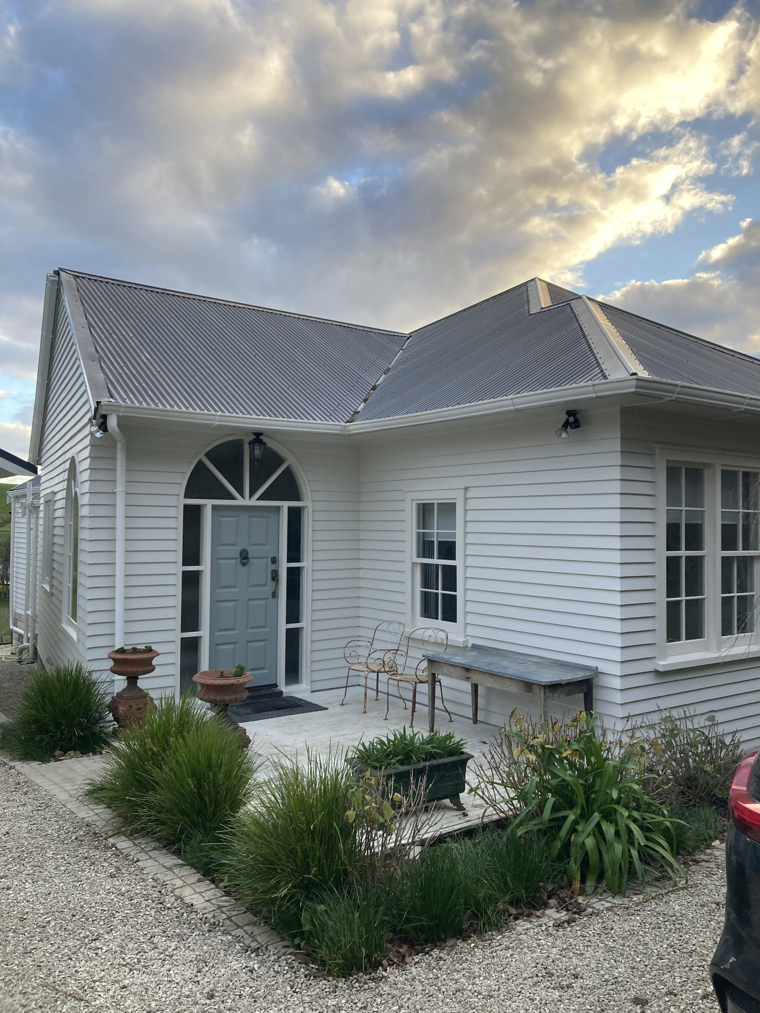 White house with light blue front door, arched window above, gray metal roof, small garden with plants and garden furniture at the entrance, cloudy sky in the background.