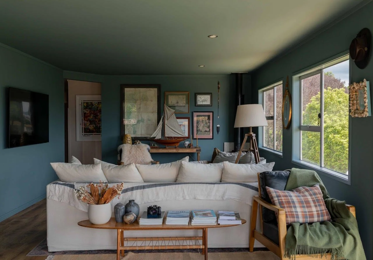 Living room with blue walls, large white sofa, armchair with plaid pillow, wooden coffee table with books and vases, framed pictures and model sailboat on shelf, multiple windows showing greenery outside, floor lamp, and wall clock.