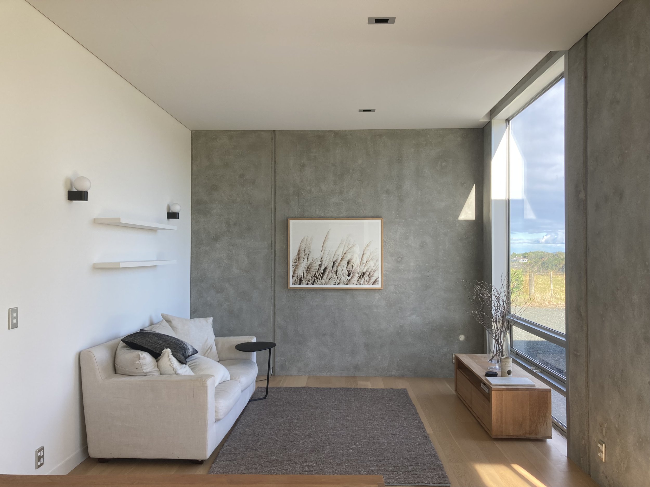 Modern living room with white sofa, black throw pillows, wall-mounted white shelves, framed artwork, a wooden coffee table with plants, large window with outdoor view, concrete and white walls, and natural light.
