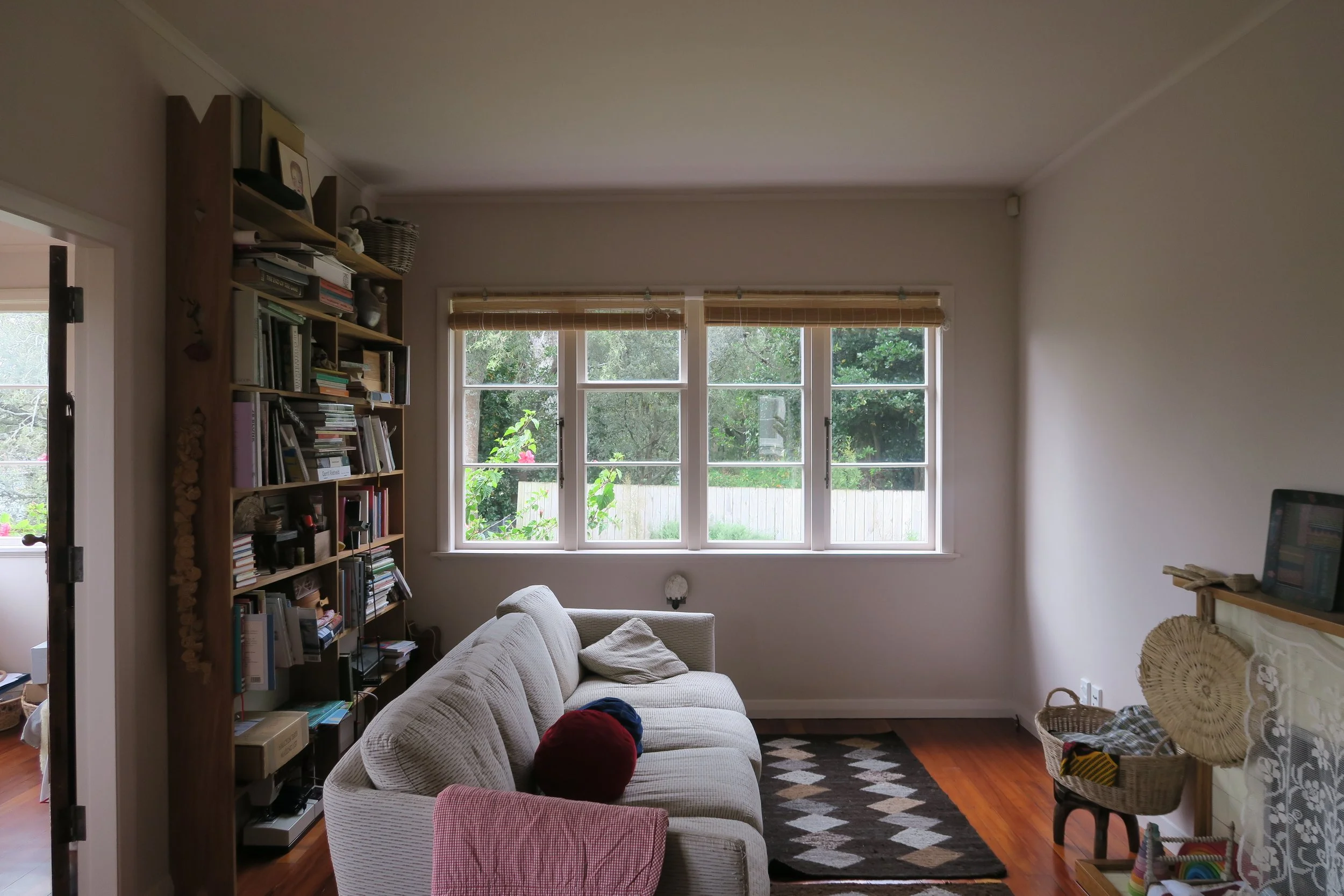 Living room with white couch, wooden bookshelf, large window facing garden, hardwood floor, small patterned rug, basket with blankets, and TV on fireplace mantel.
