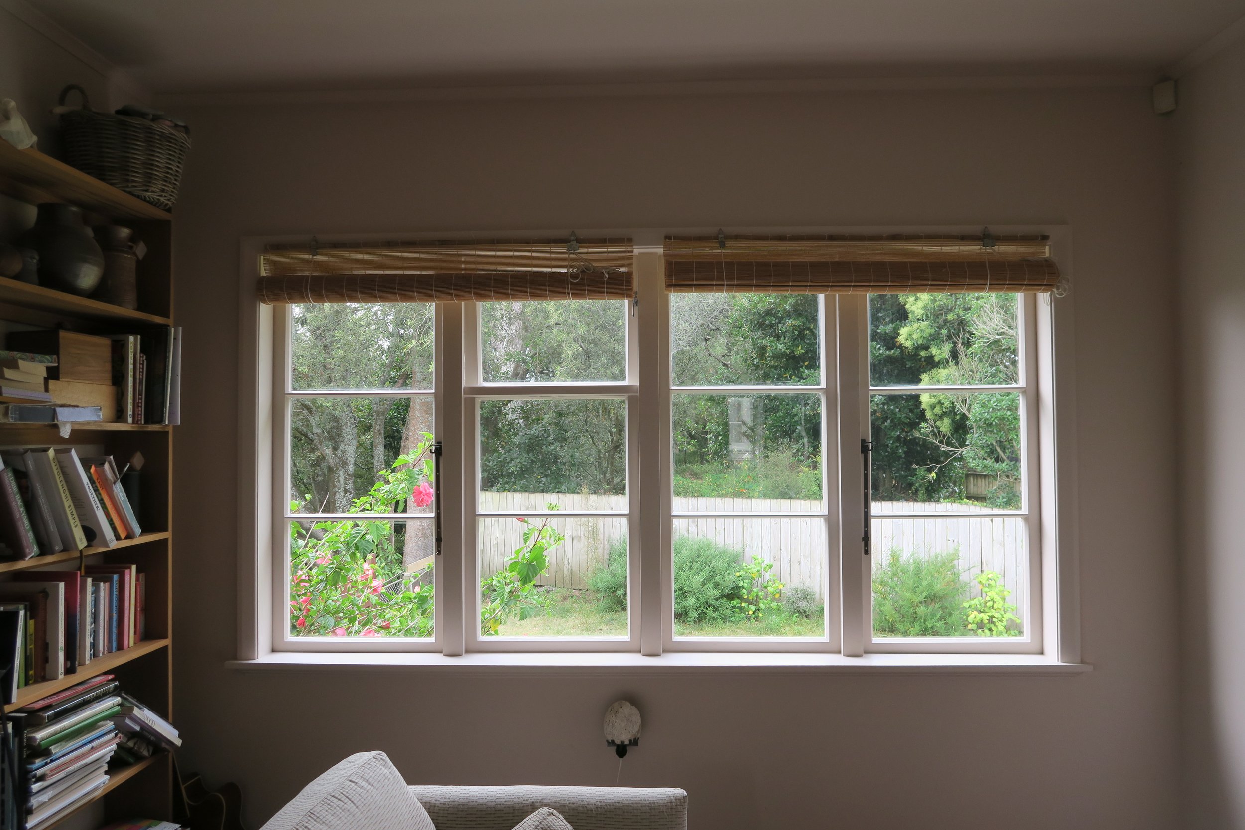Interior view of a room with a large window showing a garden and trees outside, a bookshelf filled with books on the left, and part of a light-colored sofa in the bottom left corner.